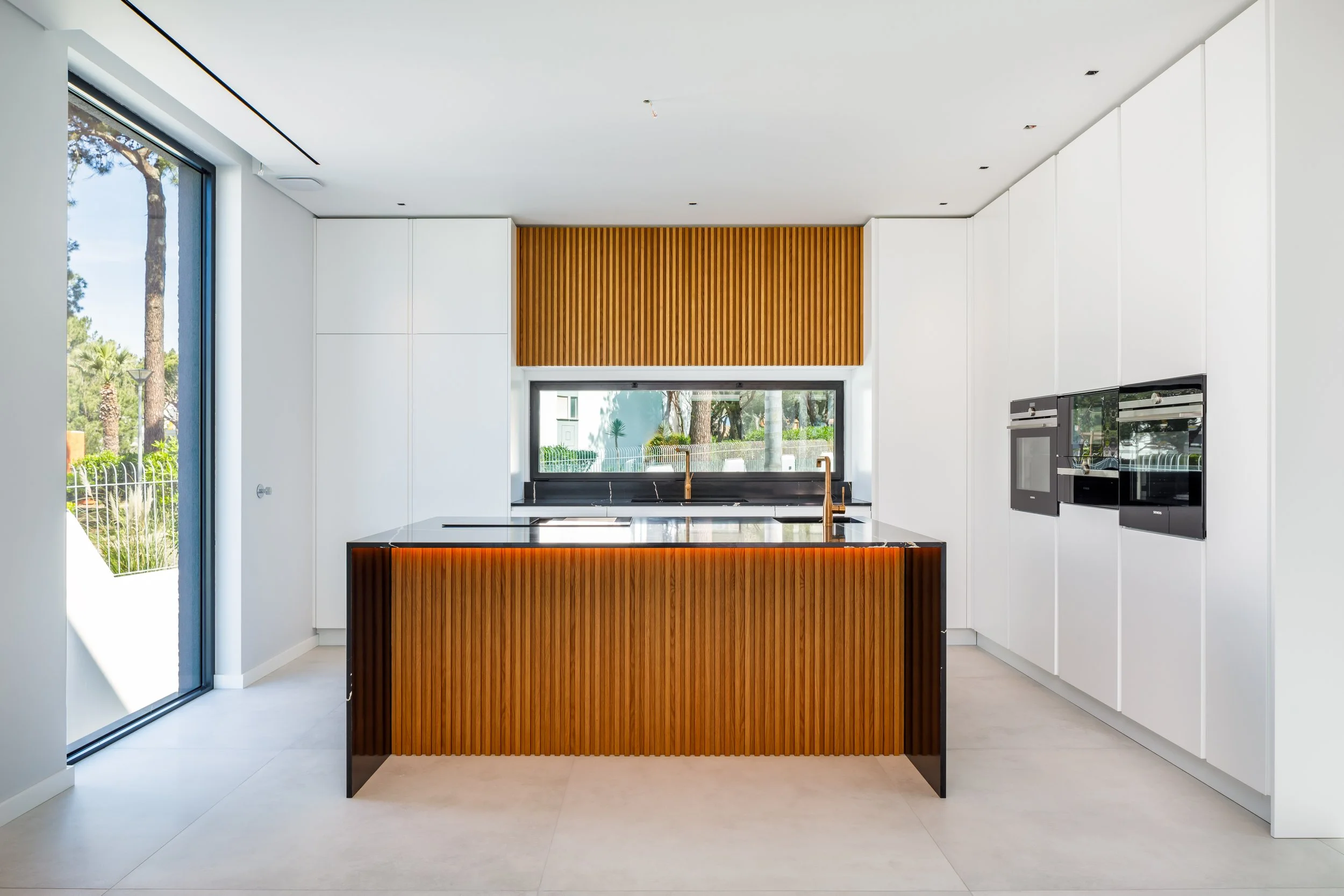 Cozinha moderna com ilha de madeira e bancada preta, paredes brancas, eletrodomésticos embutidos, janela grande atrás da pia e porta de vidro à esquerda, com vista para jardim com árvores.
