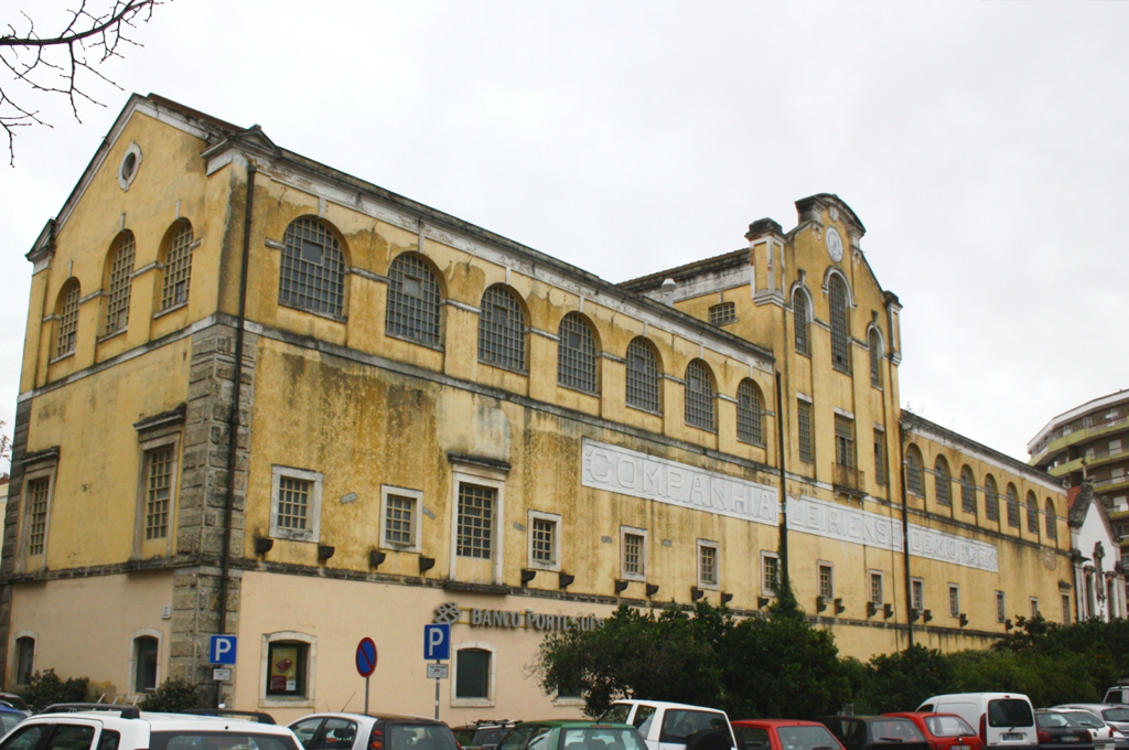 Prédio antigo de cor amarela com janelas de grade e uma placa na parede, carros estacionados na frente, árvores e céu nublado.