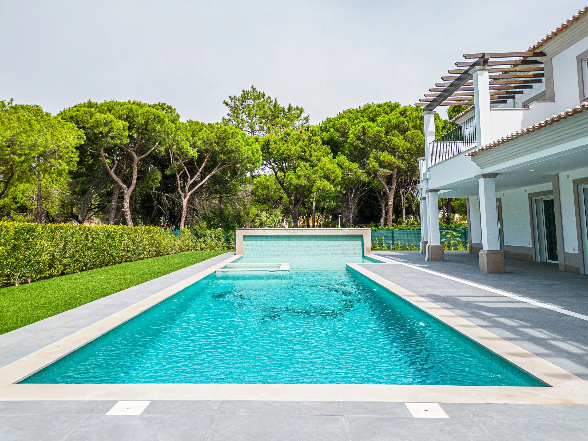 Piscina ao ar livre ao lado de uma casa moderna com varanda, cercada por árvores verdes e jardim bem cuidado.