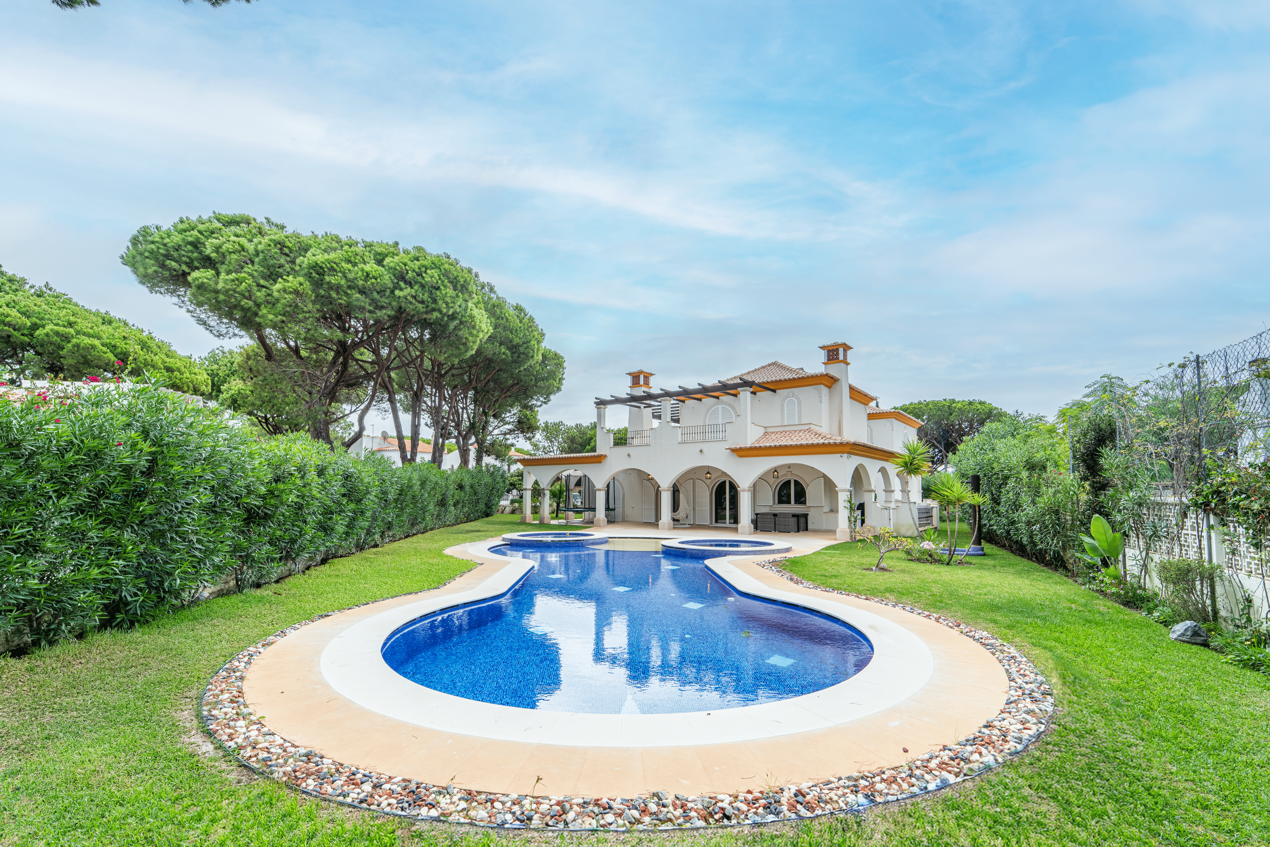 Casa branca com piscina em forma de cobra ao redor, cercada por jardim verde, árvores e ciprestes, sob céu parcialmente nublado.