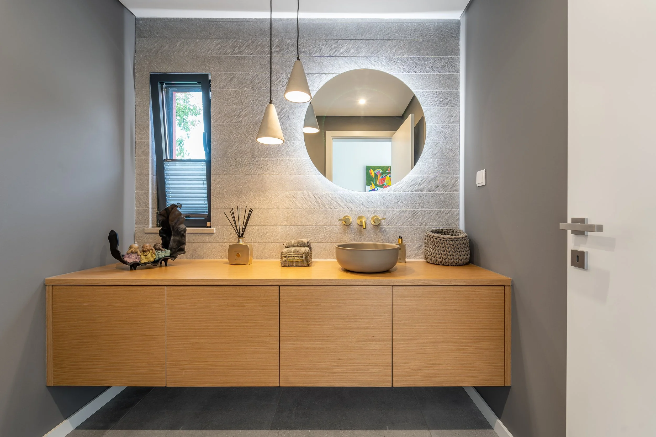 Banheiro com espelho redondo acima de uma bancada de madeira, pia redonda de cerâmica, e objetos decorativos, janela ao lado com vista para o exterior.