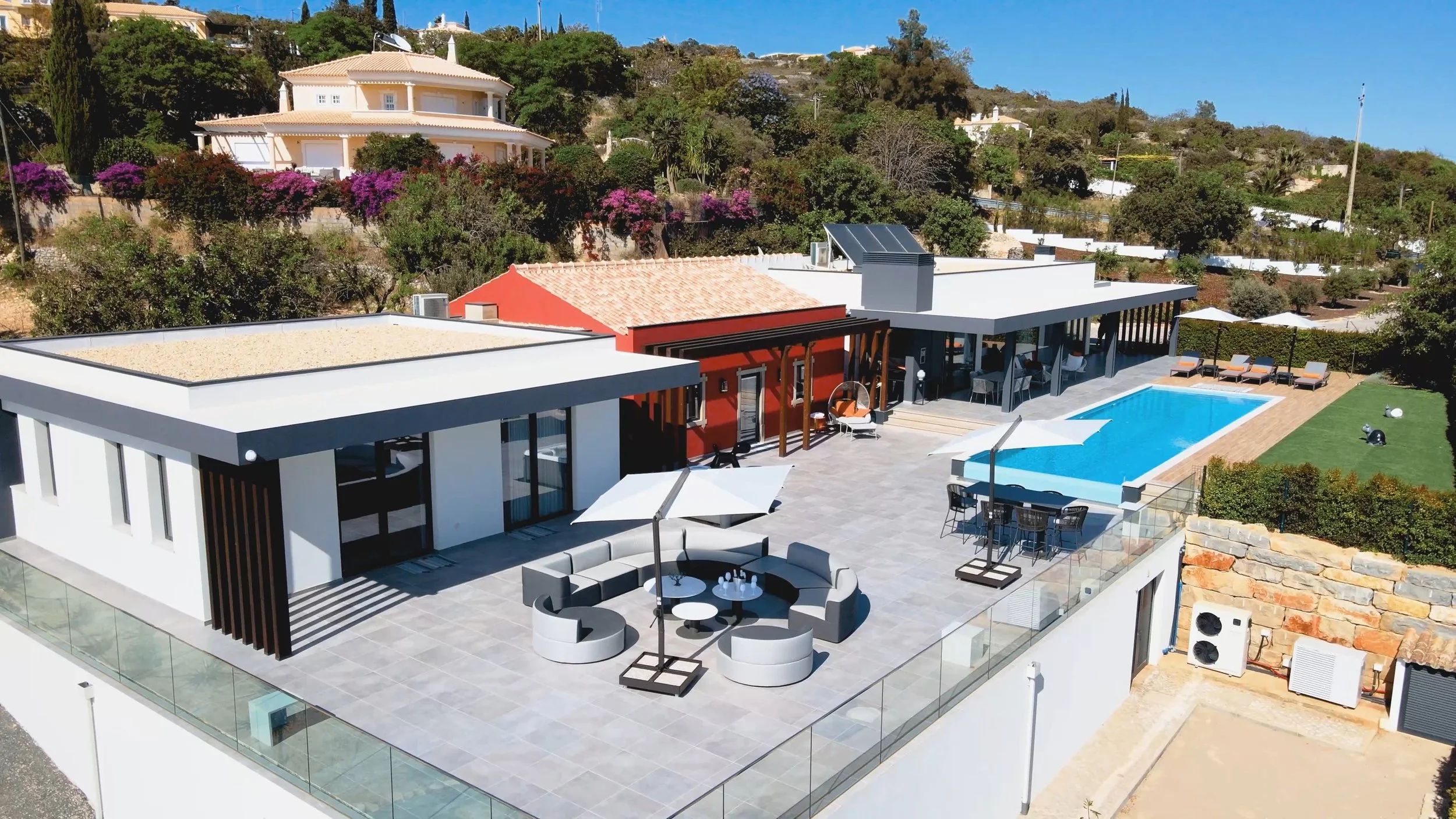 Vista aérea de uma casa moderna com área de lazer que inclui piscina, espaço com espreguiçadeiras, mesa com cadeiras e guarda-sol, em uma área com jardim e vegetação.