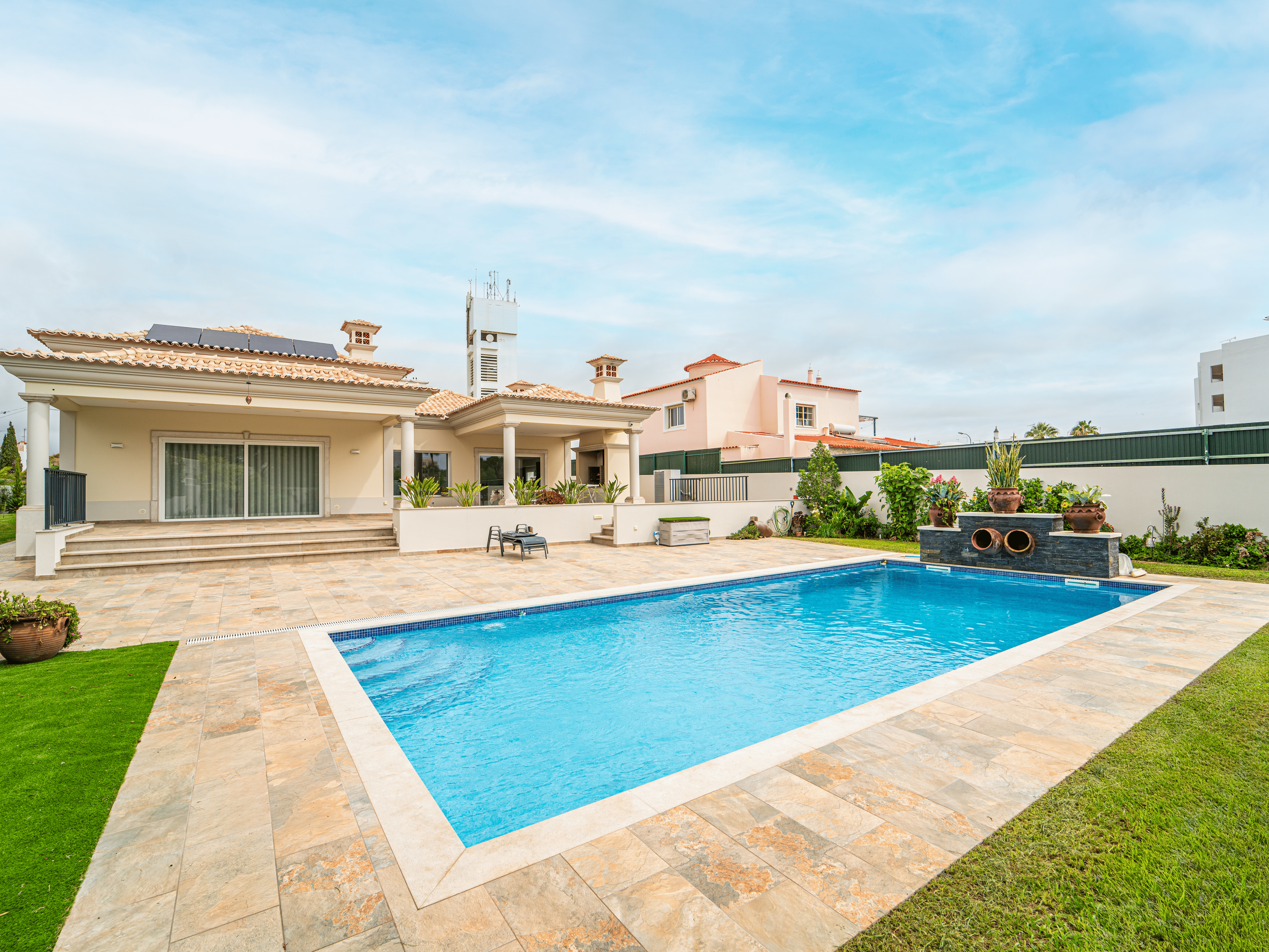 Piscina azul com zona de lazer ao redor, jardim com plantas, Casa com varanda em fundo, céu parcialmente nublado.