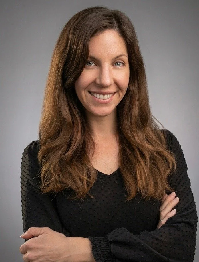 Portrait of a woman with long brown hair smiling, wearing a black textured top, crossing her arms, against a gray background.