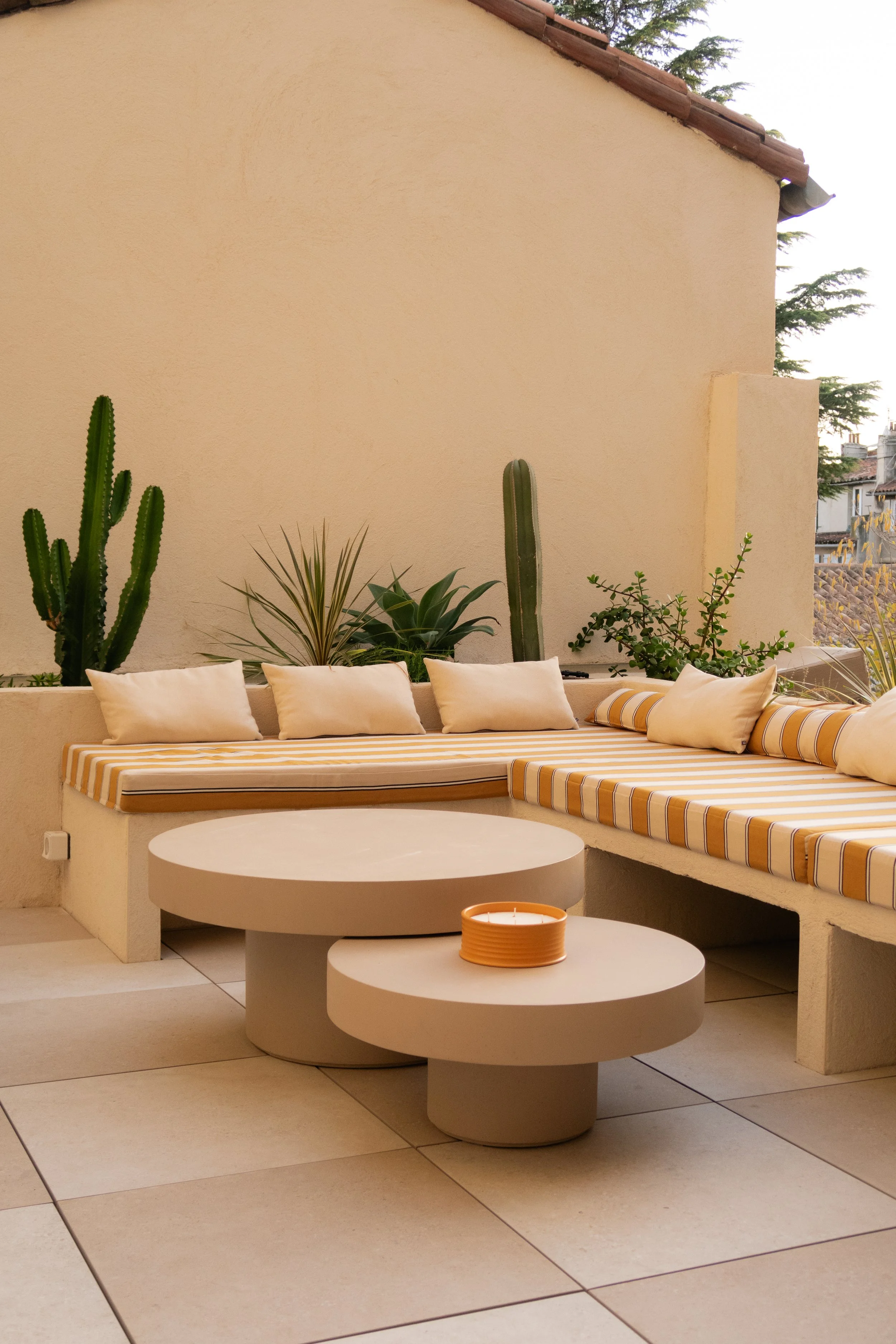 Balcon avec banquette en béton, coussins blancs et coussins rayés jaunes et blancs, table basse ronde en béton, plantes succulentes et cactus, mur beige, sol en carrelage beige.