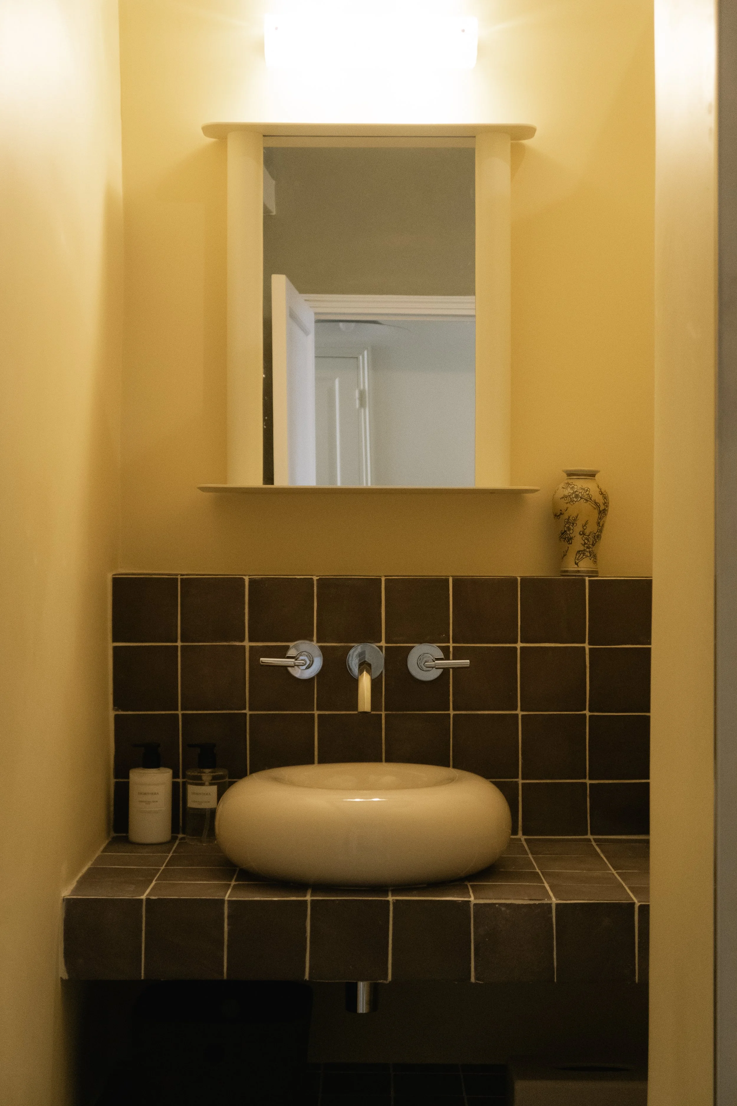 Petit lavabo ovale blanc sous un miroir, avec deux robinets en métal, dans une salle de bain avec des carrelages noirs et une petite lampe au plafond, et un vase décoratif sur une étagère.