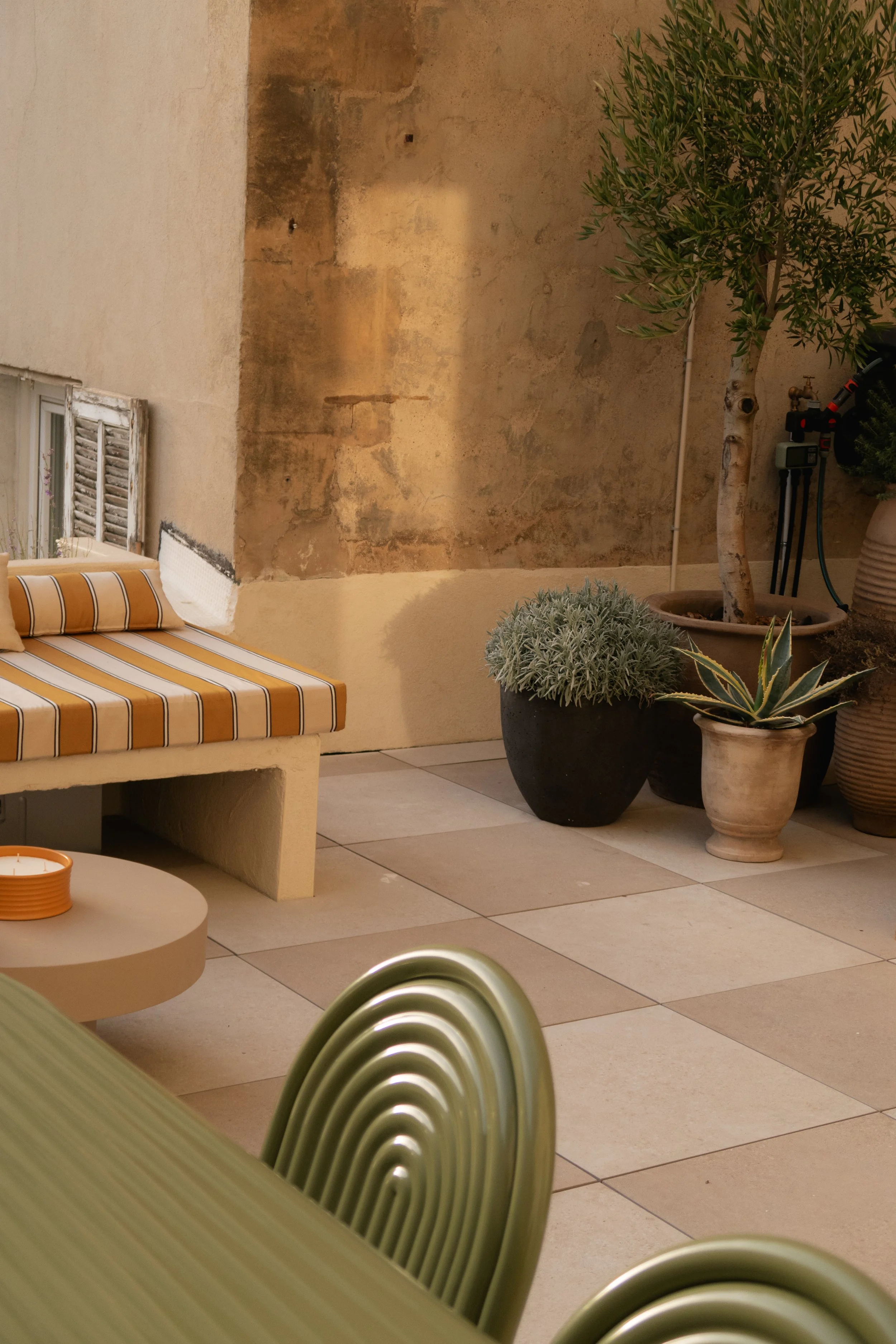 Une terrasse intérieure avec un canapé en béton, des plantes en pots, un mur en pierre, et une table verte en métal.