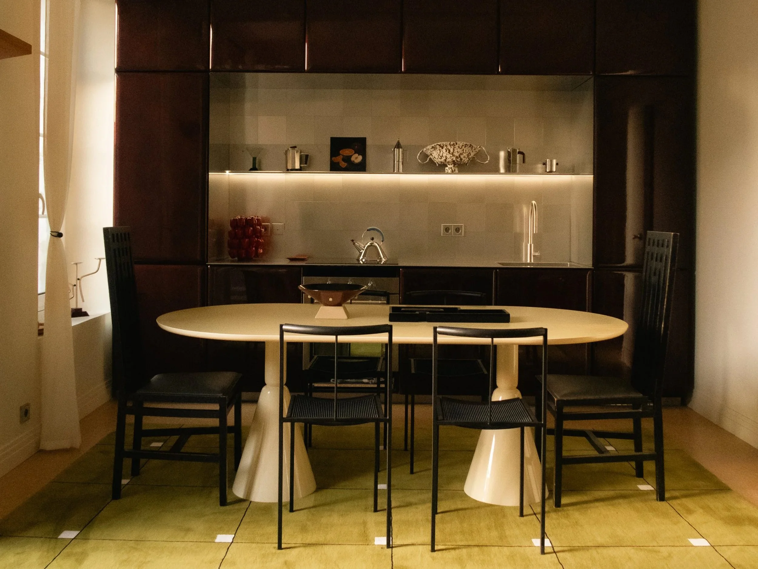 Une salle à manger avec une table ronde beige, entourée de quatre chaises noires, dans une pièce bien éclairée, avec un mur de cuisine en arrière-plan.