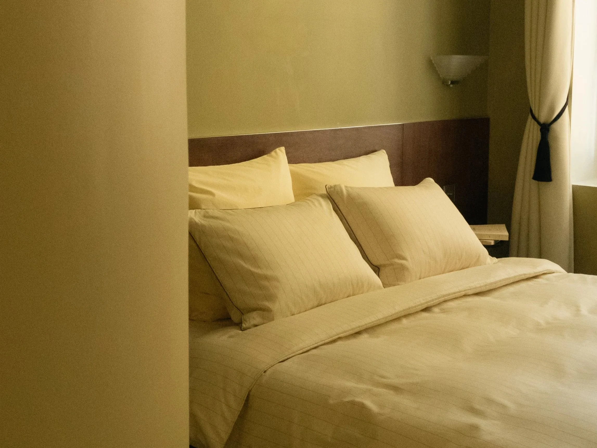 Un lit avec des oreillers beige dans une chambre d'hôtel, fenêtre avec rideaux blancs.