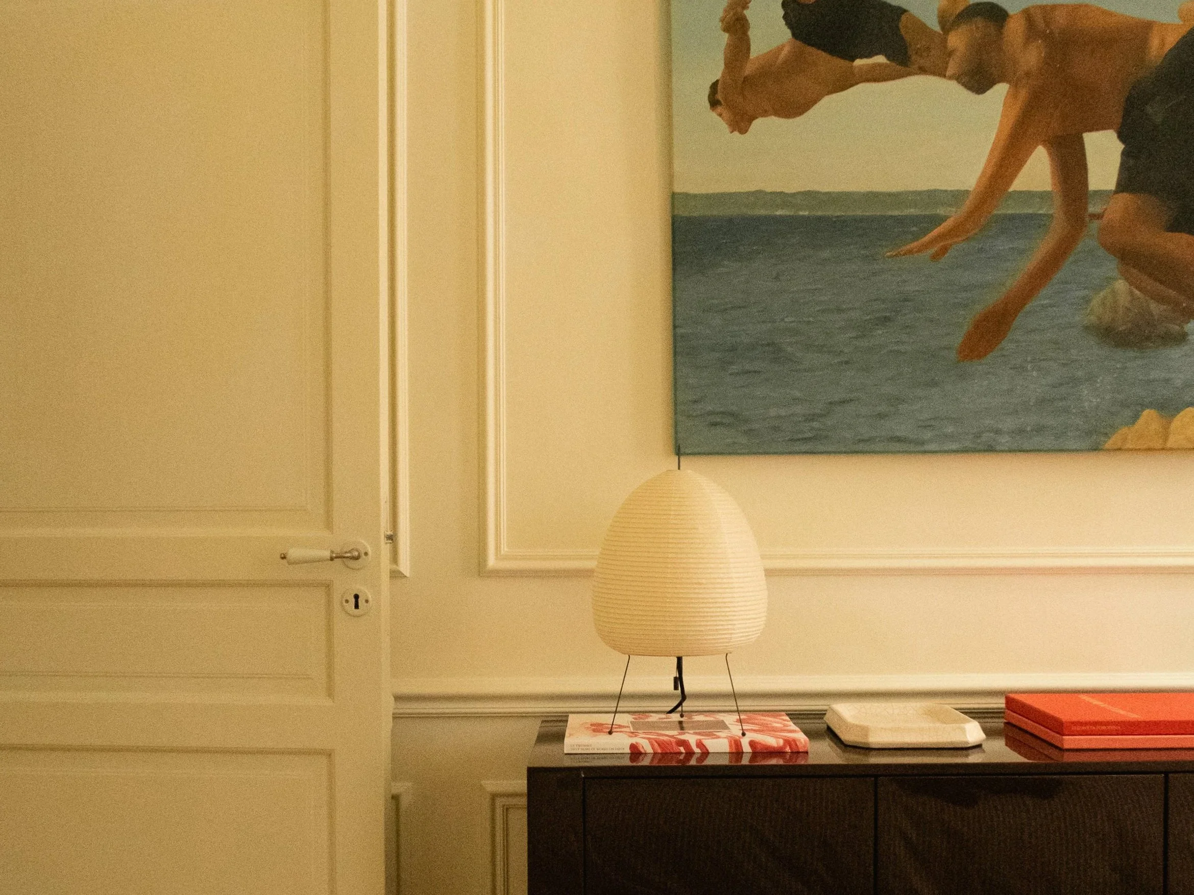Vaste tableau représentant deux hommes nageant dans la mer, avec un ciel clair en arrière-plan, accroché au mur au-dessus d'une console noire avec une lampe en forme de boule en papier et quelques objets sur la surface.