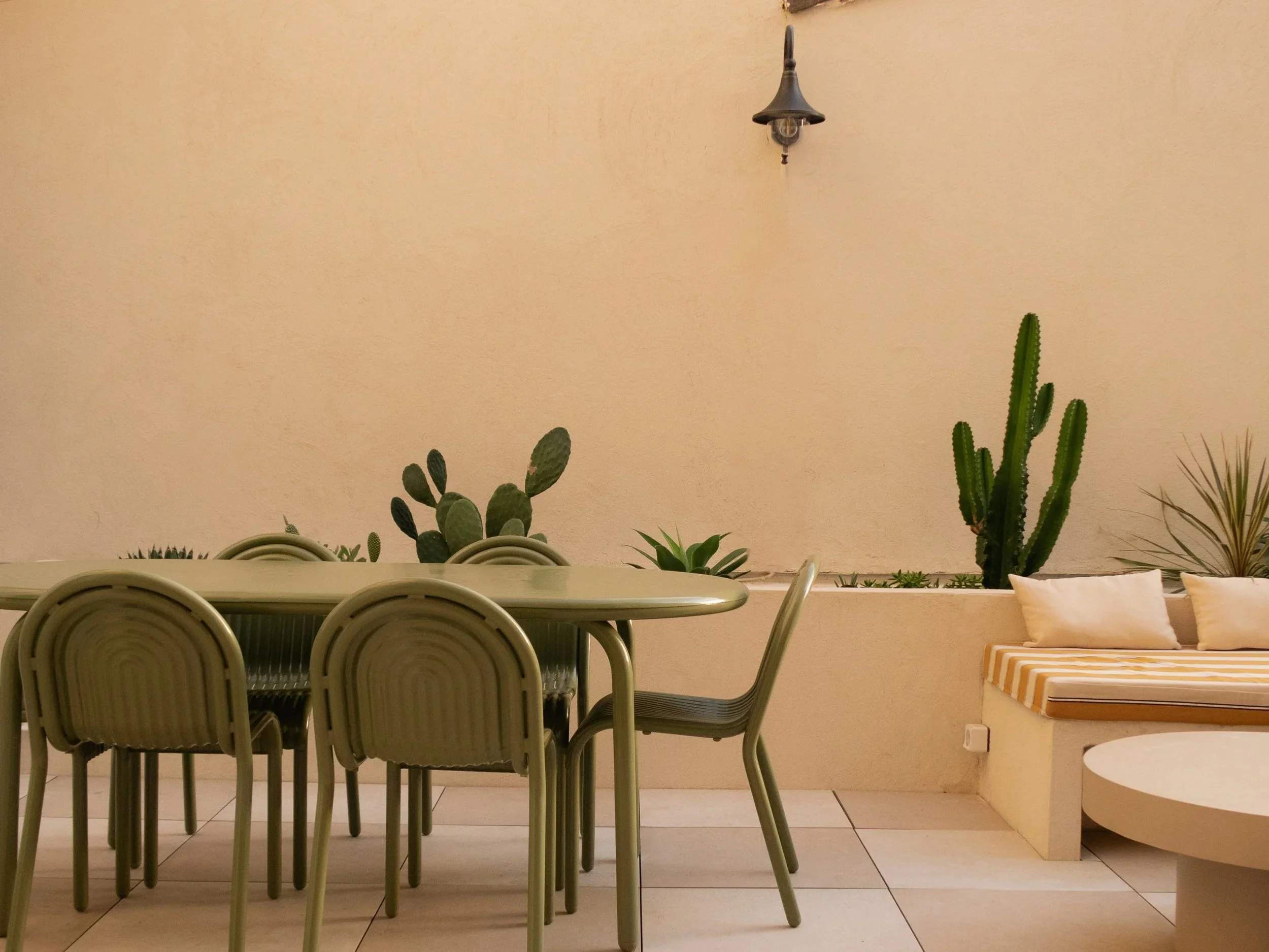 Espace extérieur avec une table et plusieurs chaises en métal vert, une banquette avec coussins, et plusieurs plantes en pot dont des cactus en arrière-plan.