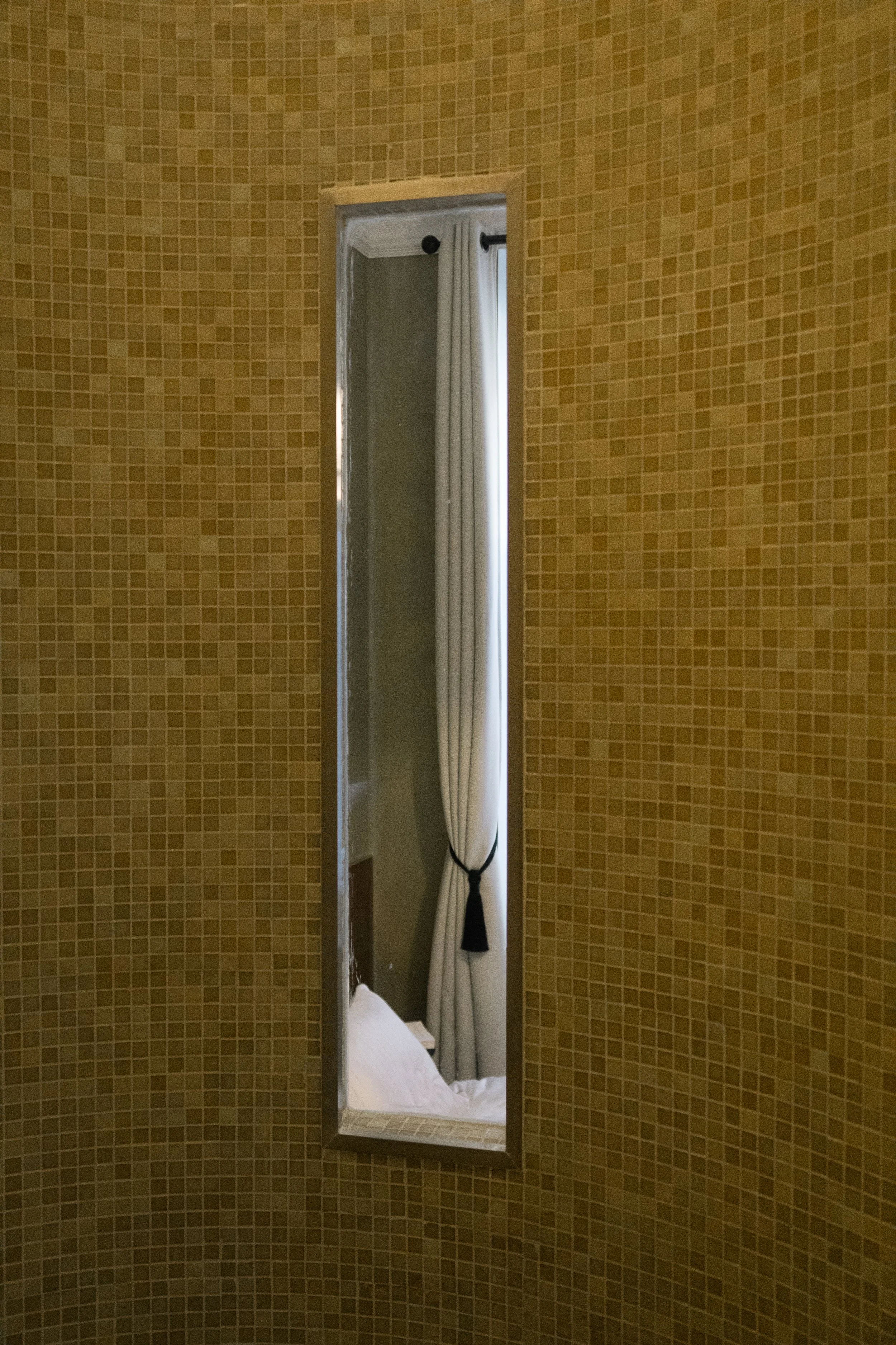 Un miroir rectangulaire fixé sur un mur en mosaïque jaune, reflétant une porte avec un rideau blanc attaché avec un ruban noir.