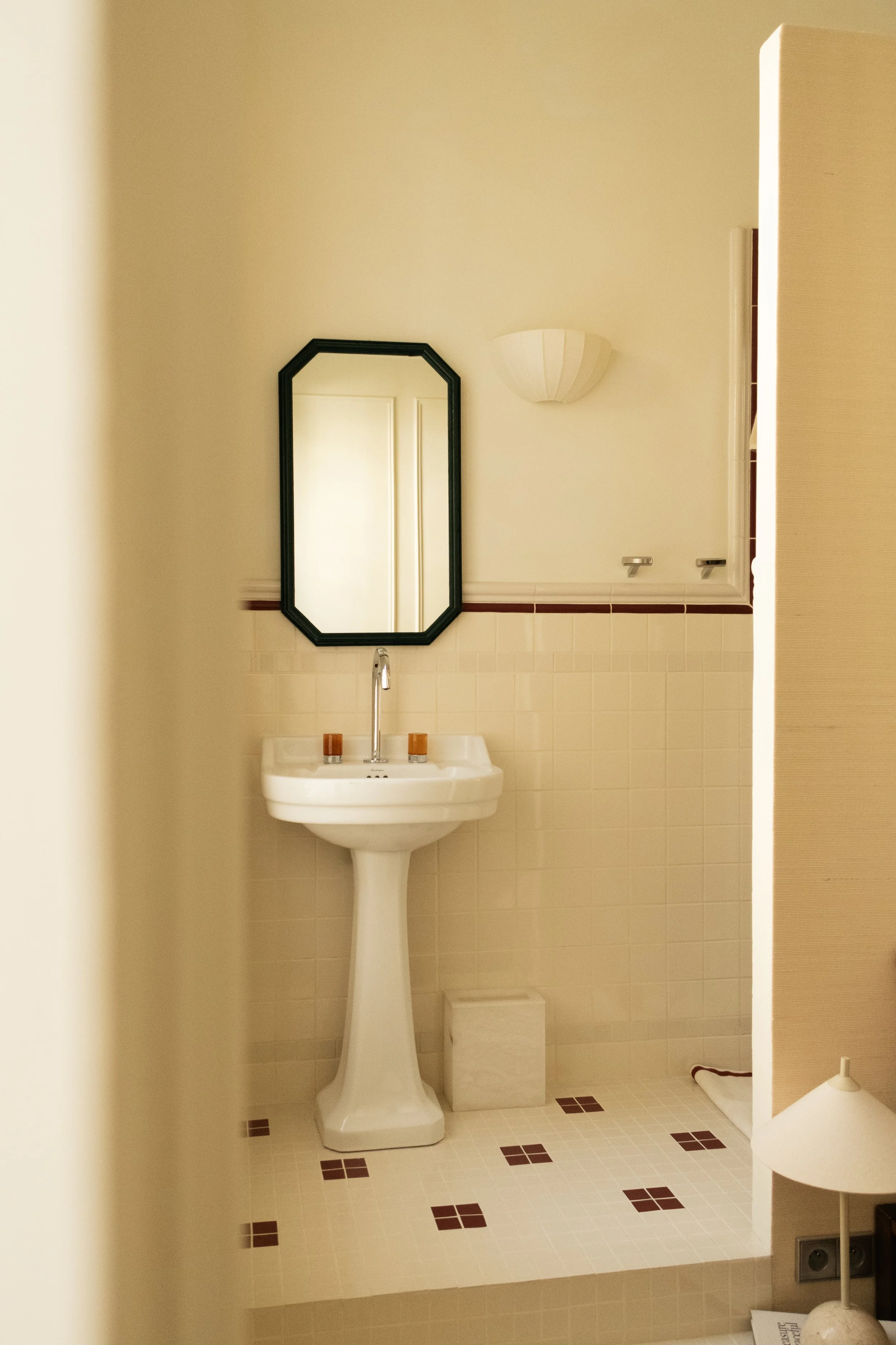 Petite salle de bain avec un lavabo blanc, un miroir rectangulaire à cadre noir, une applique murale blanche, un miroir de pharmacie à gauche et une petite lampe de table à droite.