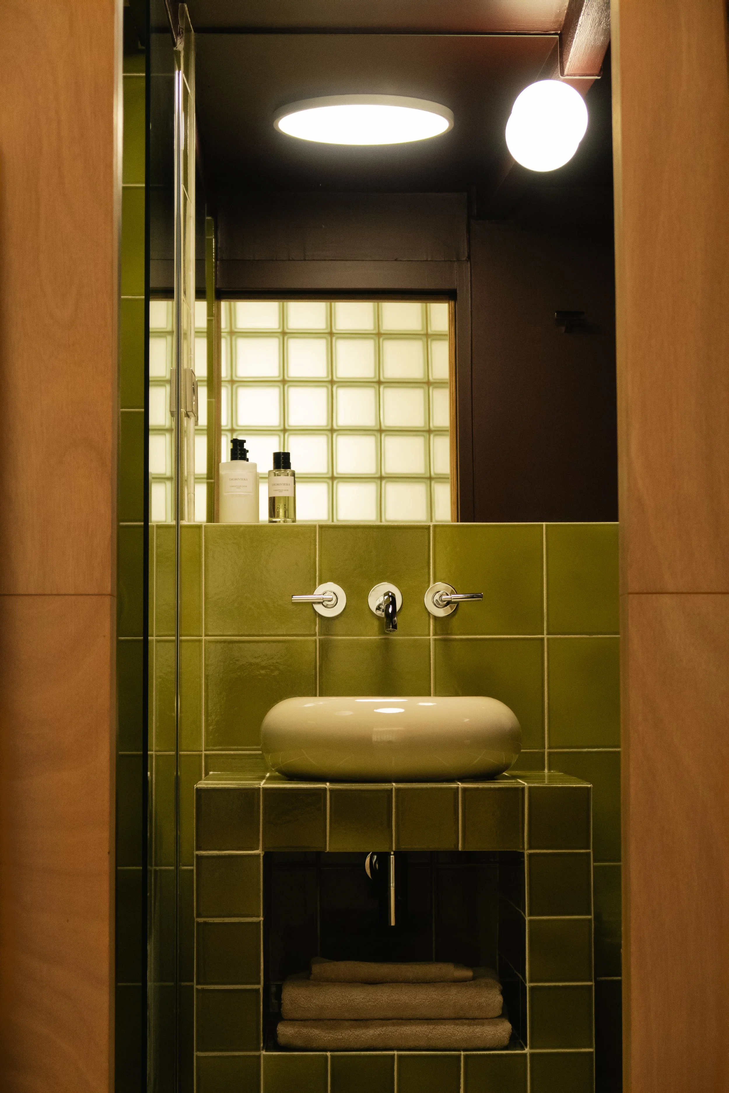 Vue d'une salle de bain avec des carreaux verts, un lavabo ovale blanc, et des bouteilles de savon ou lotion sur le rebord. Miroir derrière, éclairage rond au plafond, et un mur en verre de blocs translucides dans le fond.