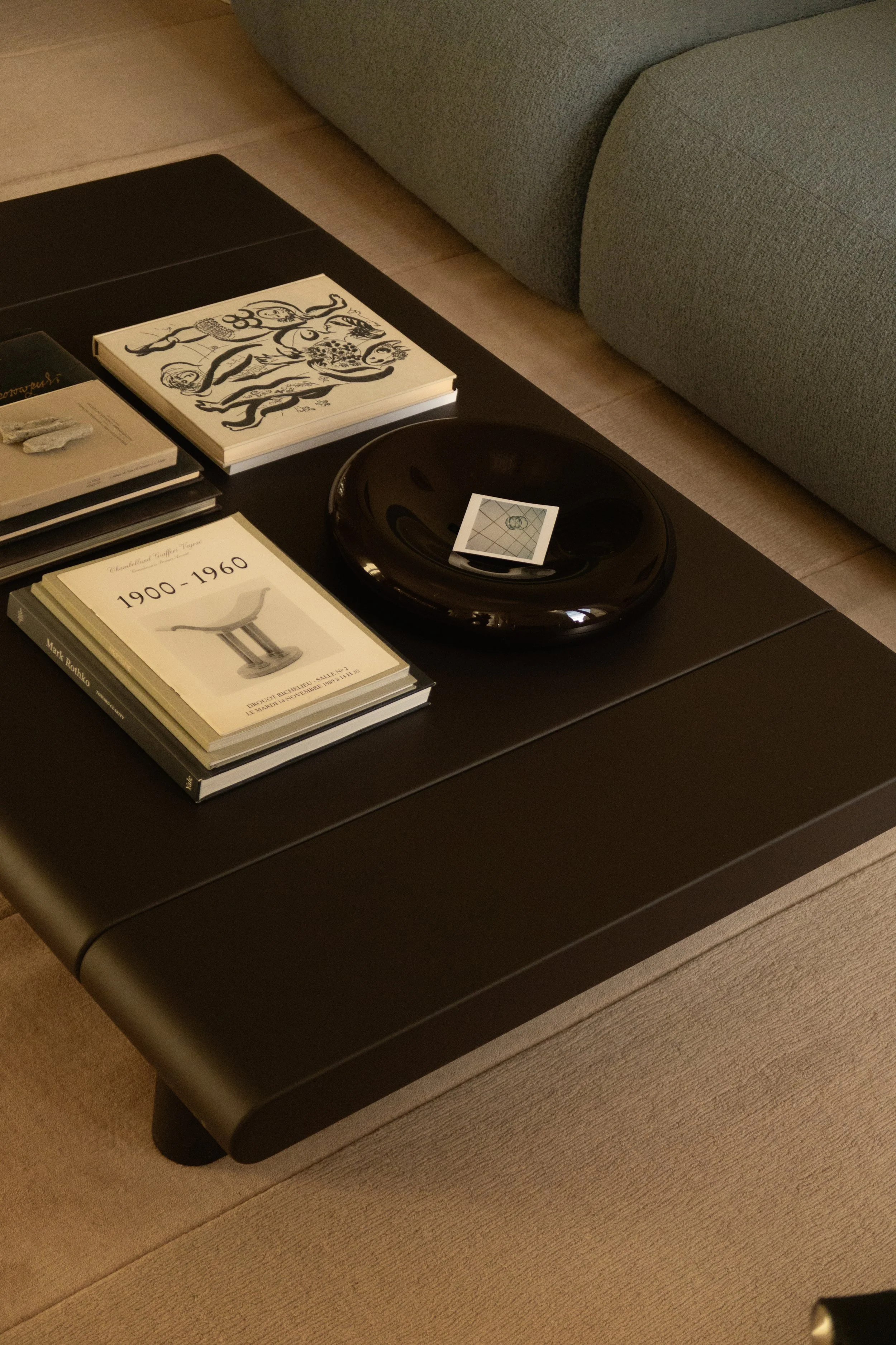 Table basse noire avec livres, une coupe en verre noir contenant une carte ou un petit objet, dans un salon avec canapé gris et tapis beige.