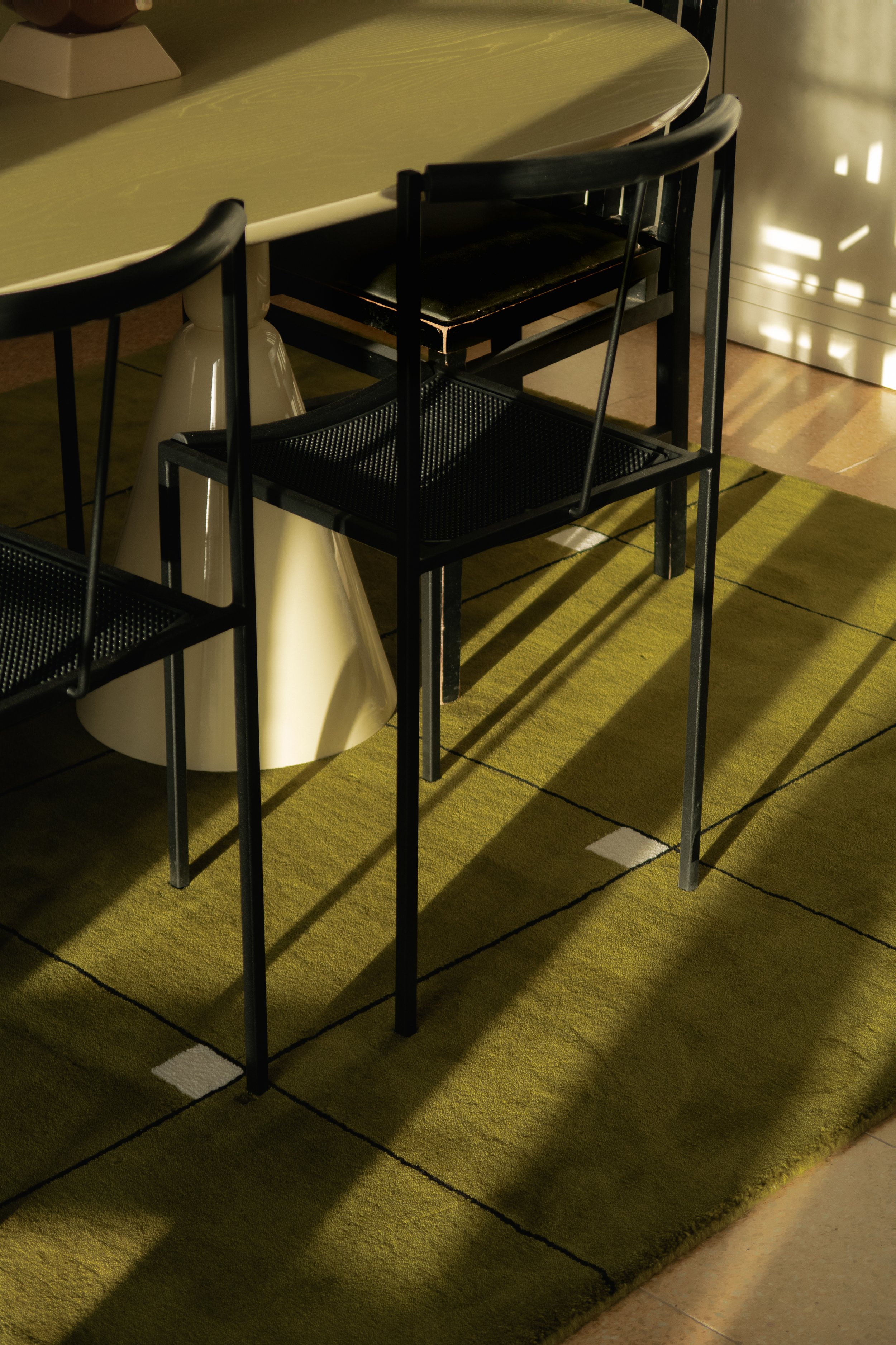 Une salle avec une table ovale beige, entourée de chaises noires, et un tapis vert avec des carrés blancs et noirs. La lumière du soleil crée des ombres sur le sol.
