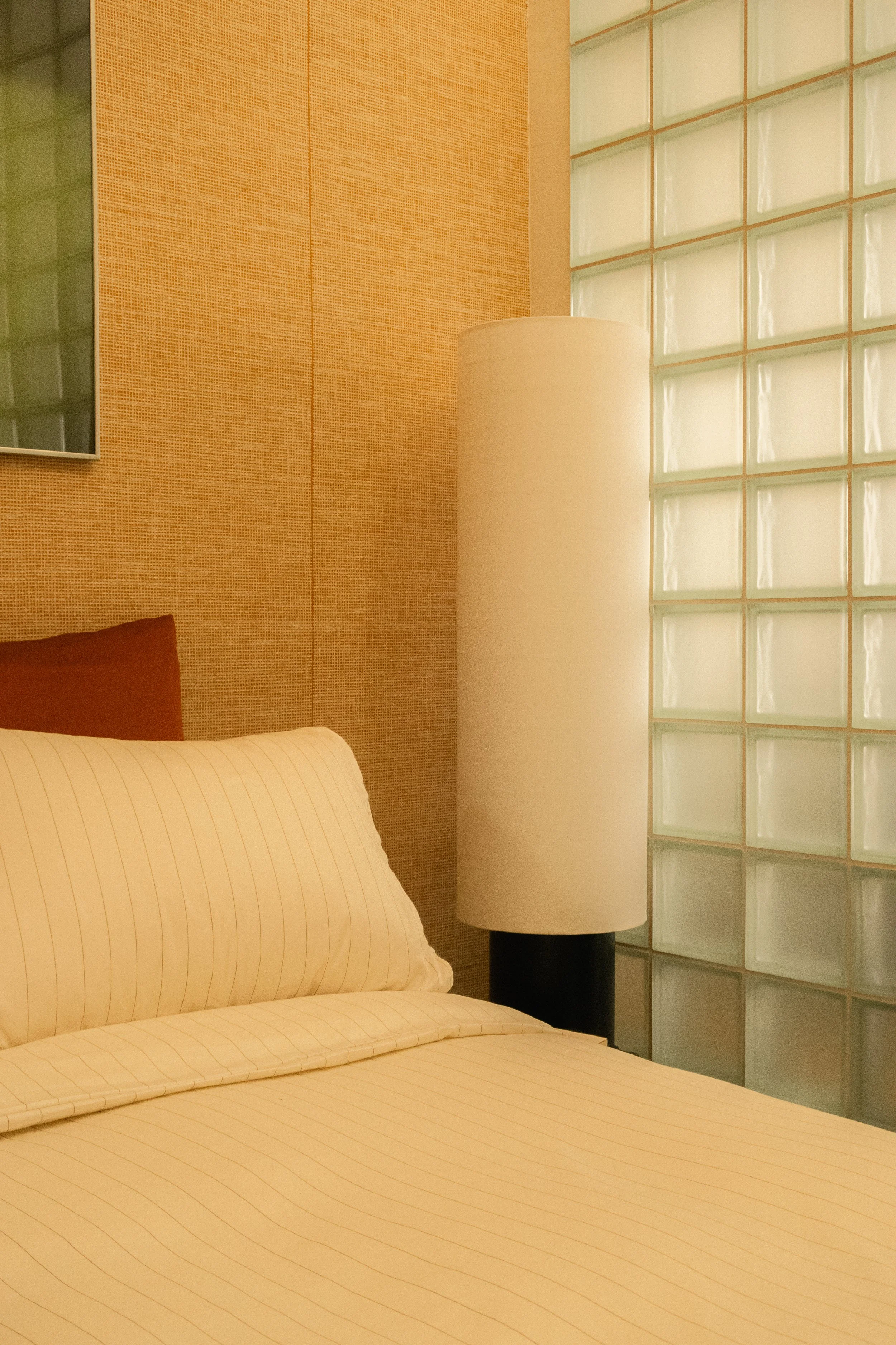 L'intérieur d'une chambre avec un lit aux draps rayés, un coussin beige, une lampe de sol blanche et un mur en briques de verre.