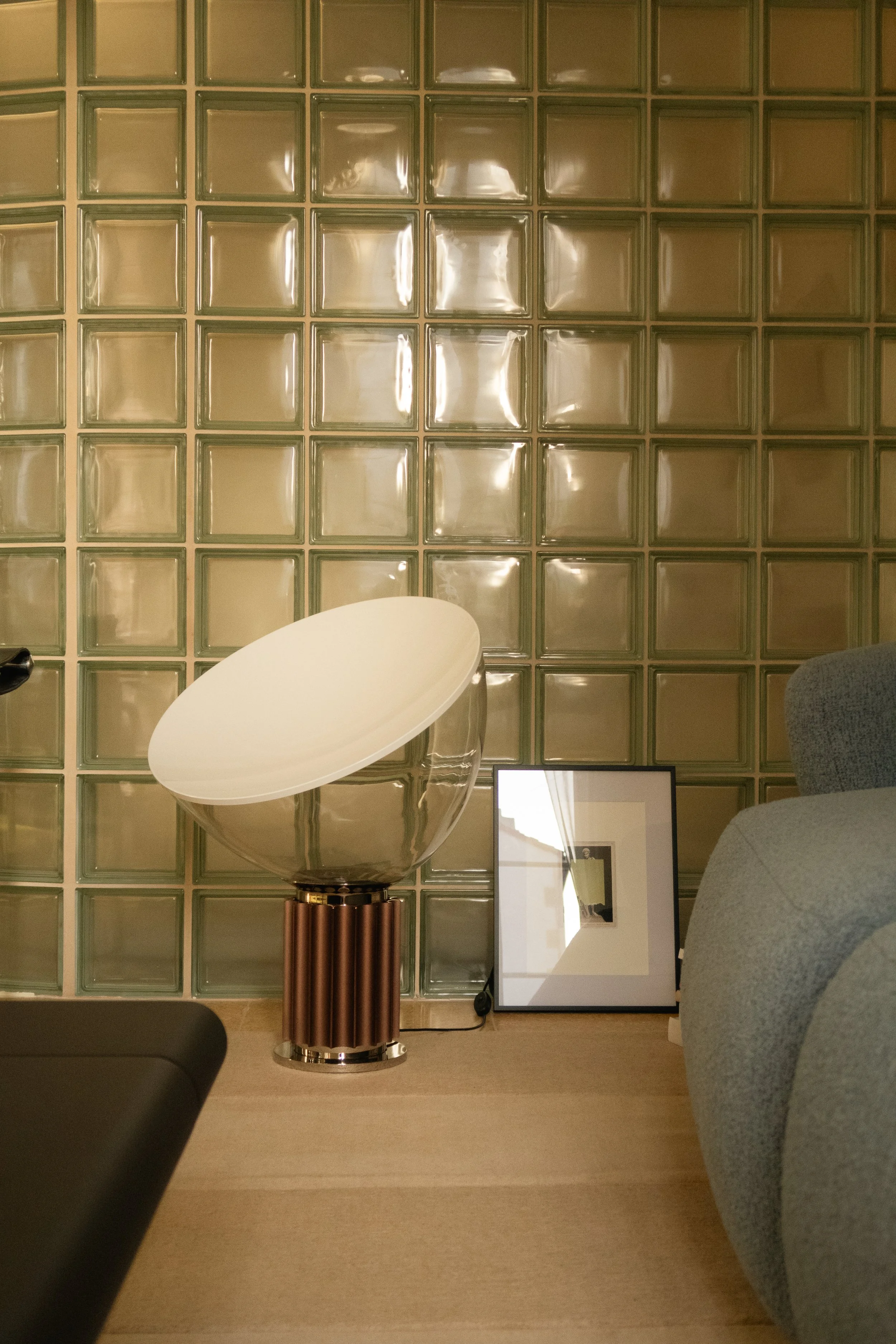 Une pièce avec un mur en blocs de verre, un fauteuil gris, une lampe de designer avec un abat-jour blanc, et un cadre photo posé sur le sol.