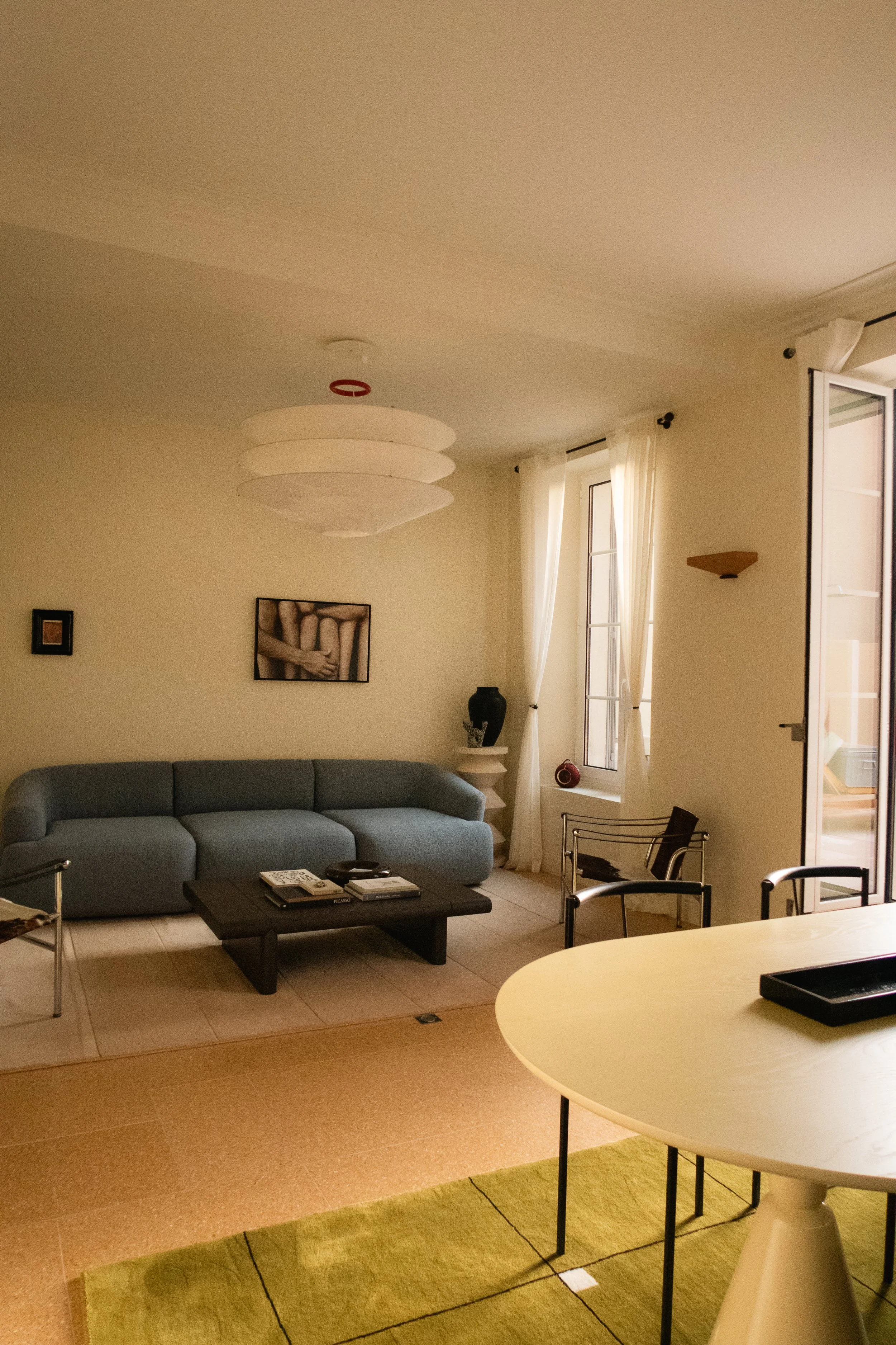 Salon avec un canapé gris, une table basse, une fenêtre avec rideaux blancs, et un éclairage tamisé.