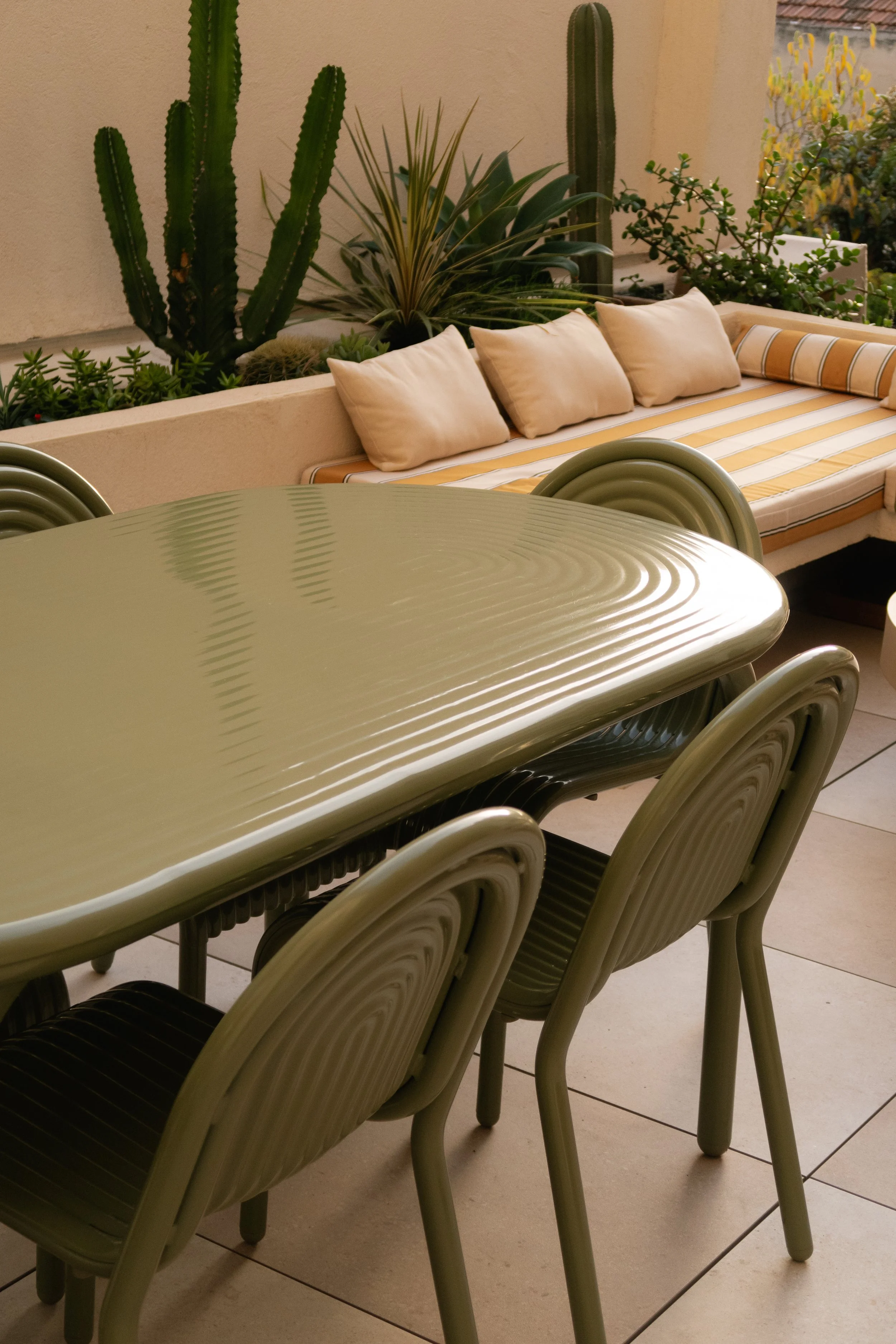 Espace extérieur avec une table et des chaises en métal vert, un banc avec des coussins et des plantes cactus en arrière-plan.