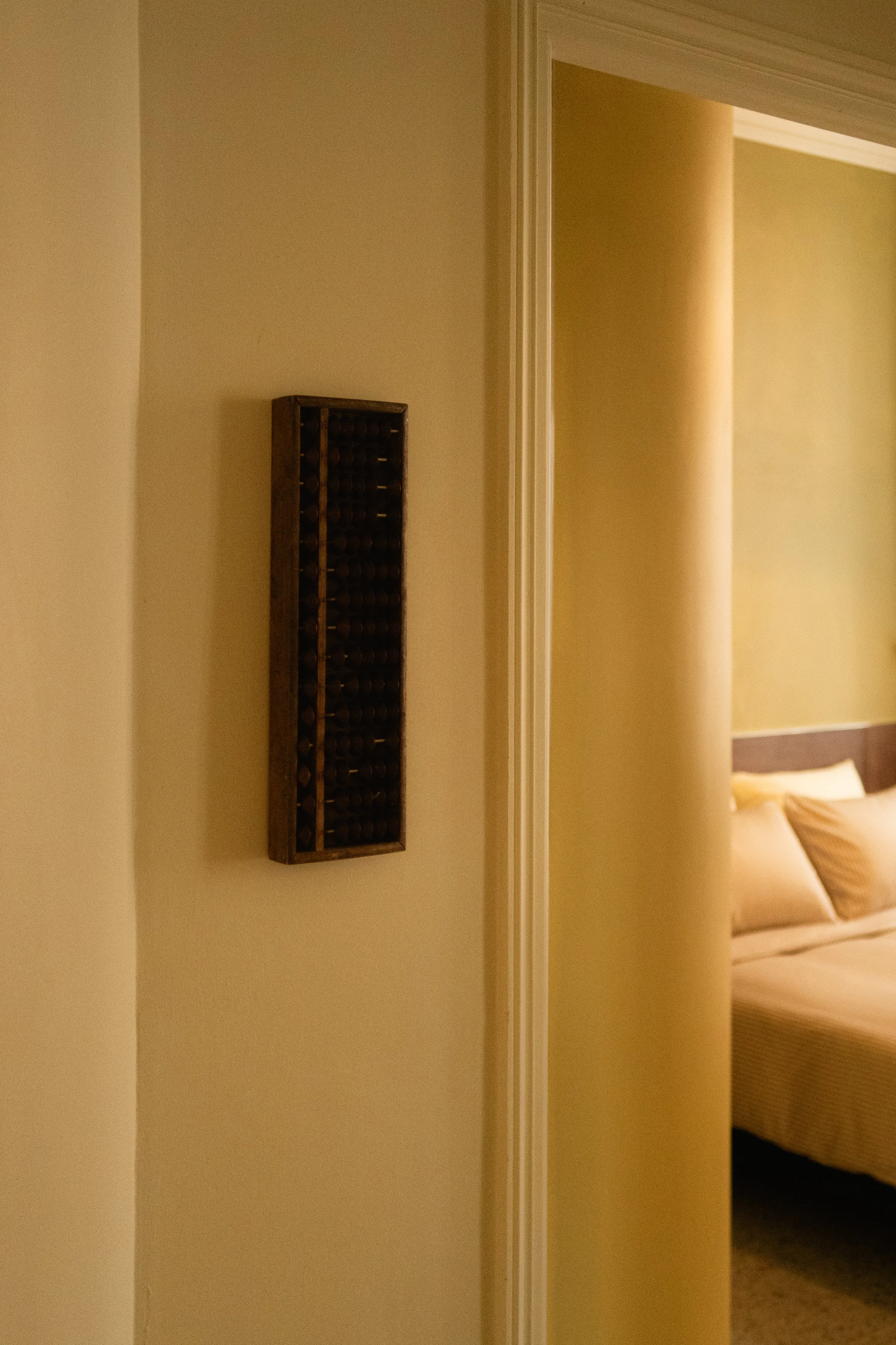 Une pièce avec un mur pâle, un petit casier à vin en bois accroché au mur, et une chambre visible à travers une porte ouverte, avec un lit à draps beige et des oreillers.