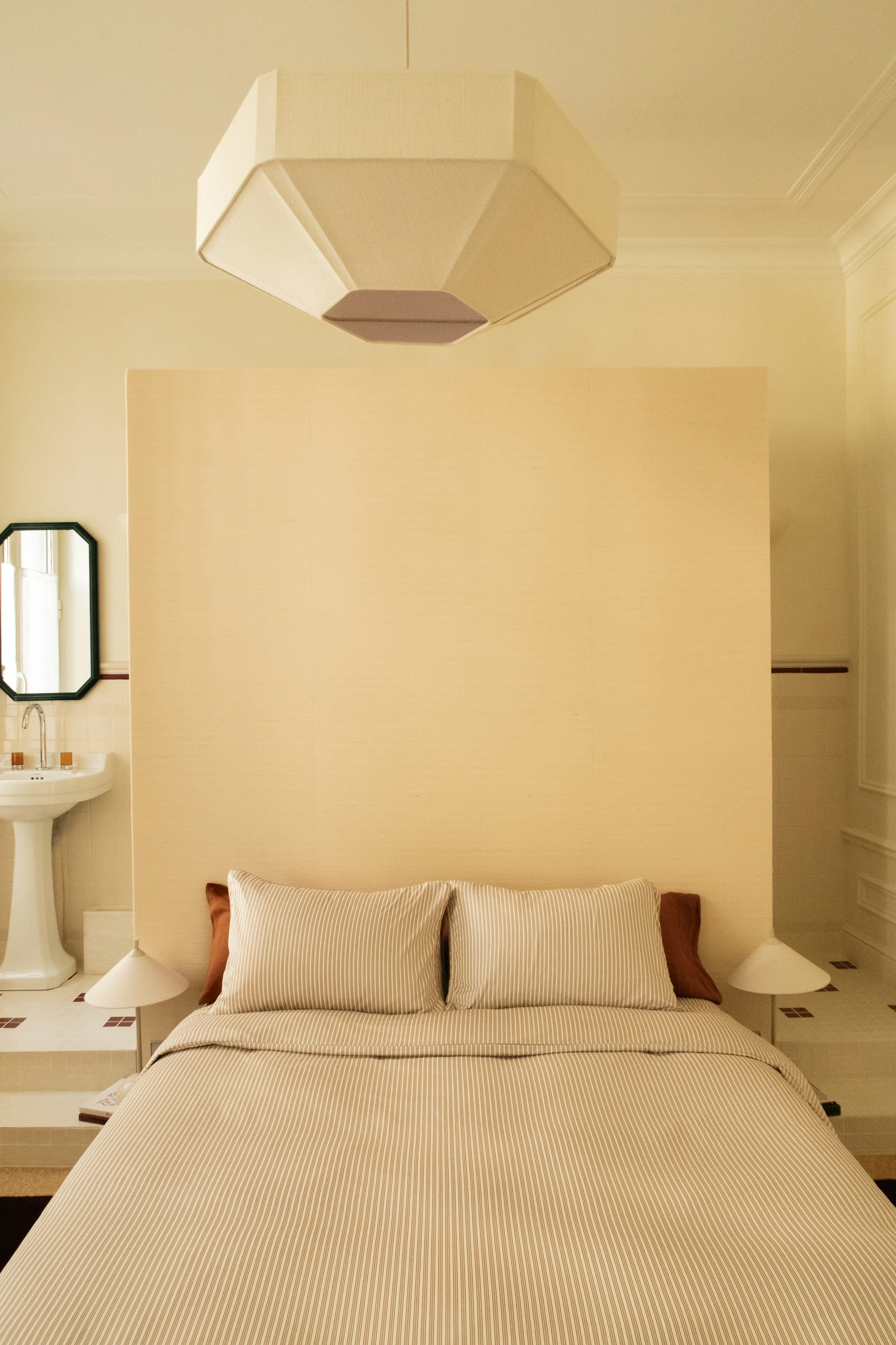 Chambre avec un lit fait avec des draps rayés, des oreillers et une tête de lit en bois clair, avec une lampe de chaque côté. En arrière-plan, un mur blanc séparant la chambre d'une petite salle de bain avec un lavabo et un miroir noir.