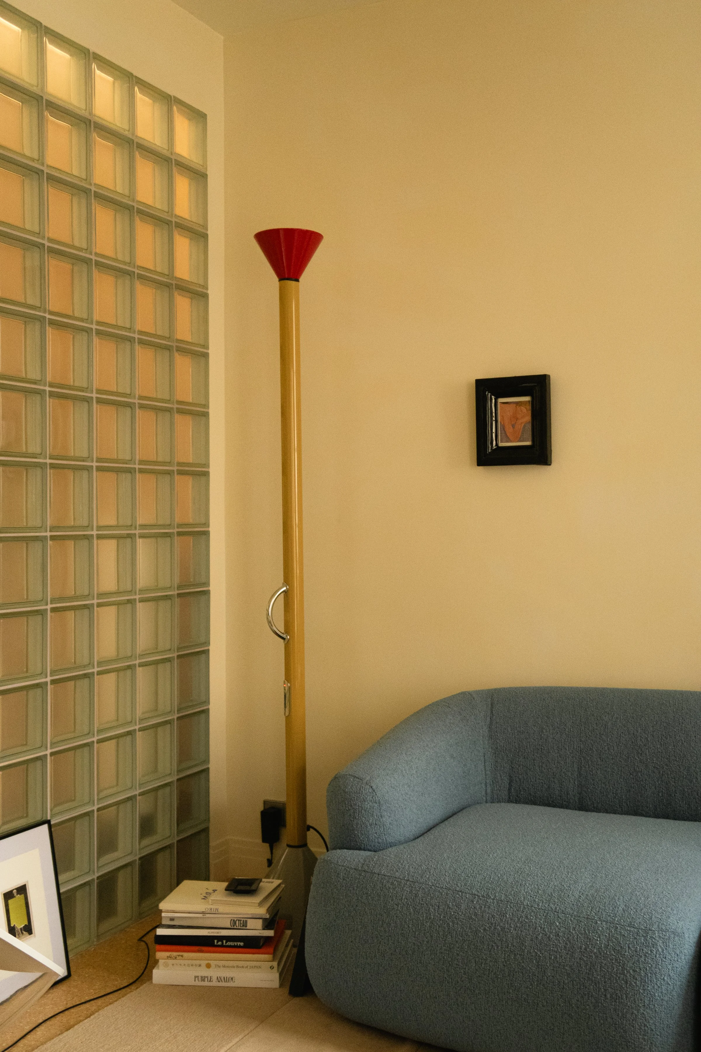 Un coin salon avec un canapé bleu, une lampe sur pied en bois avec un abat-jour rouge, une pile de livres sur le sol et une petite œuvre d'art encadrée sur le mur, avec un mur en blocs de verre à gauche.