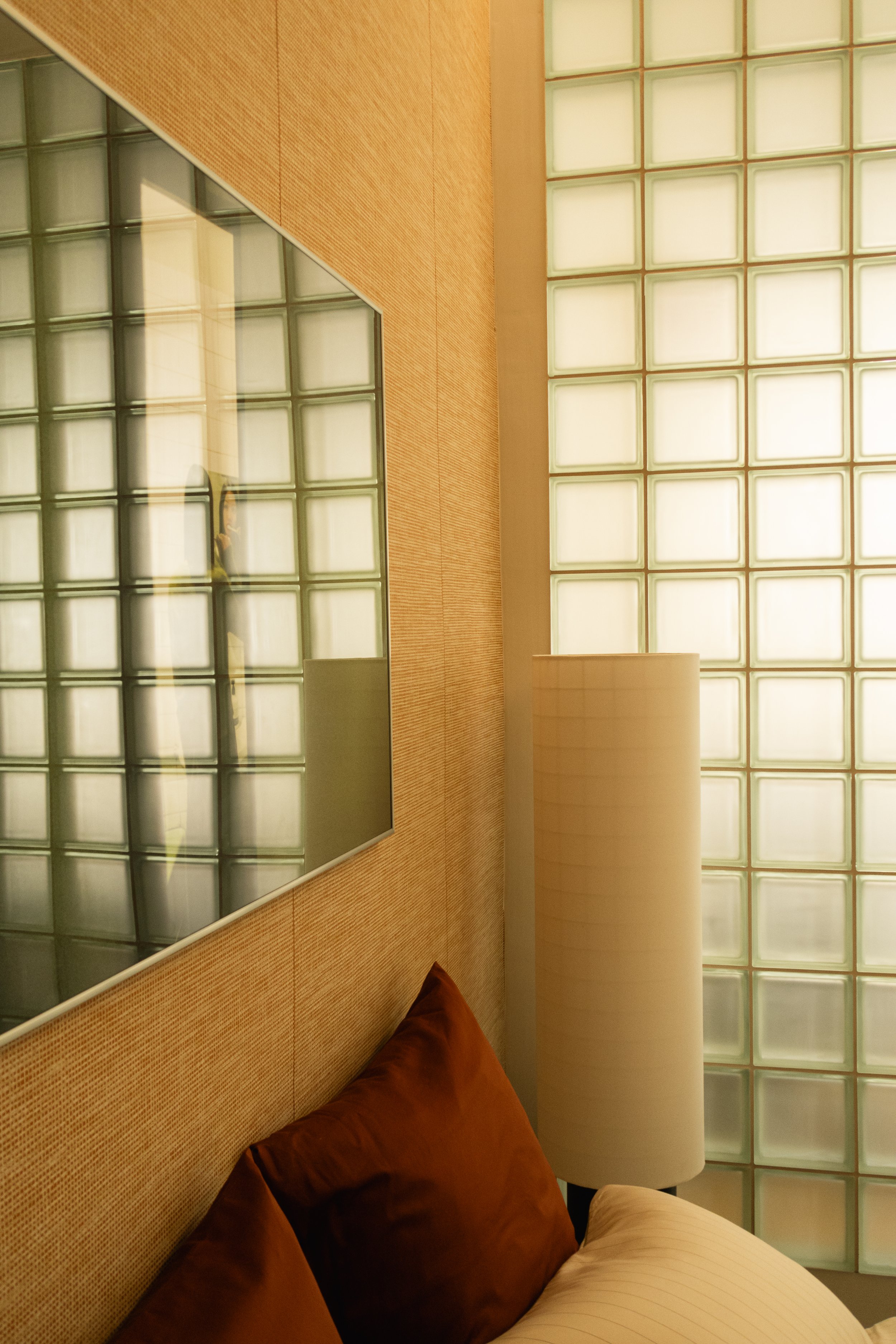Un coin de chambre avec un lit, des coussins marron, un miroir rectangulaire, un lampadaire blanc, un mur en verre cassettes et un mur en bois.