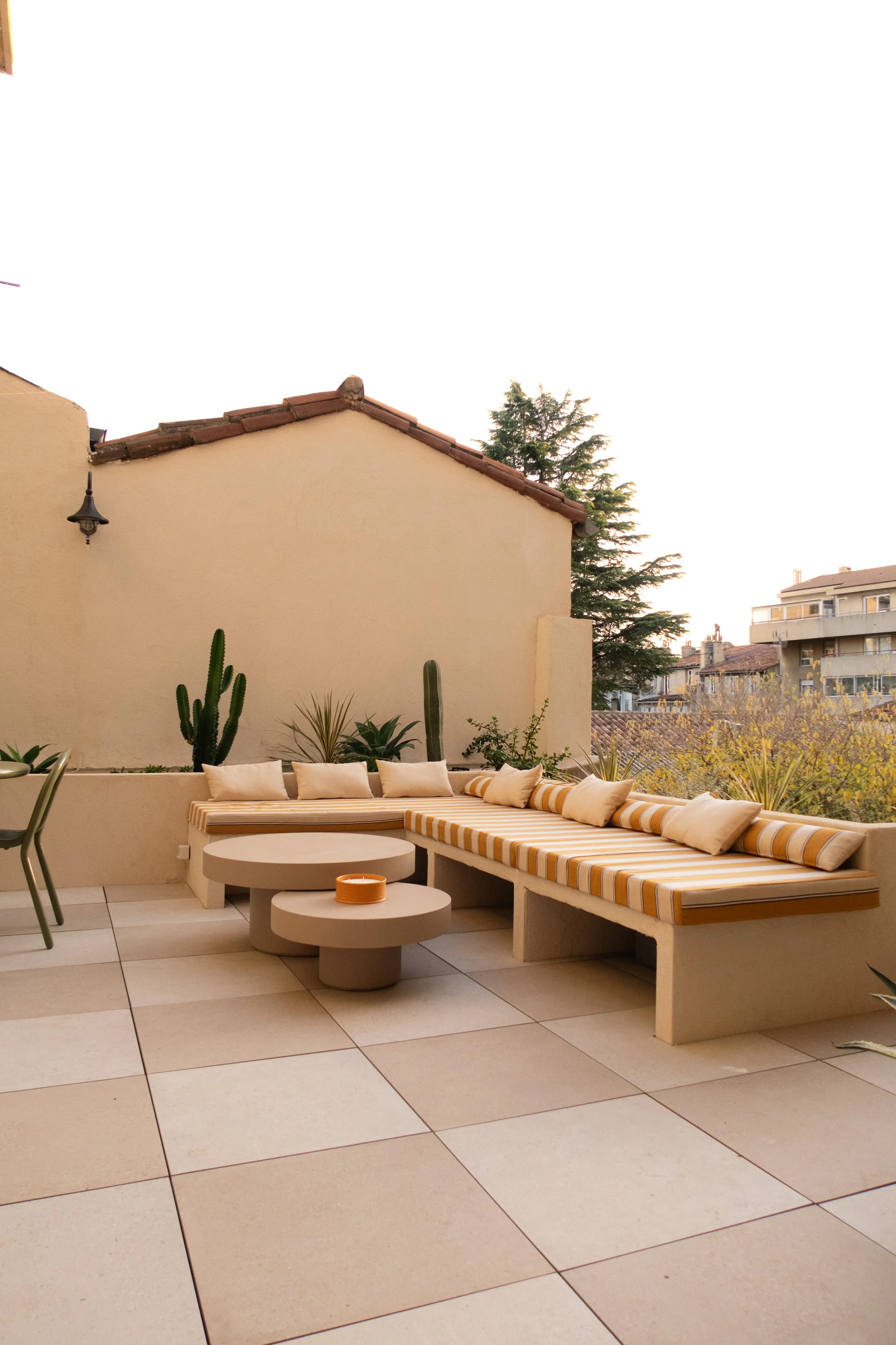 Terrasse avec mobilier moderne, coussins et plantes cactées, vue sur des bâtiments et des arbres en arrière-plan.