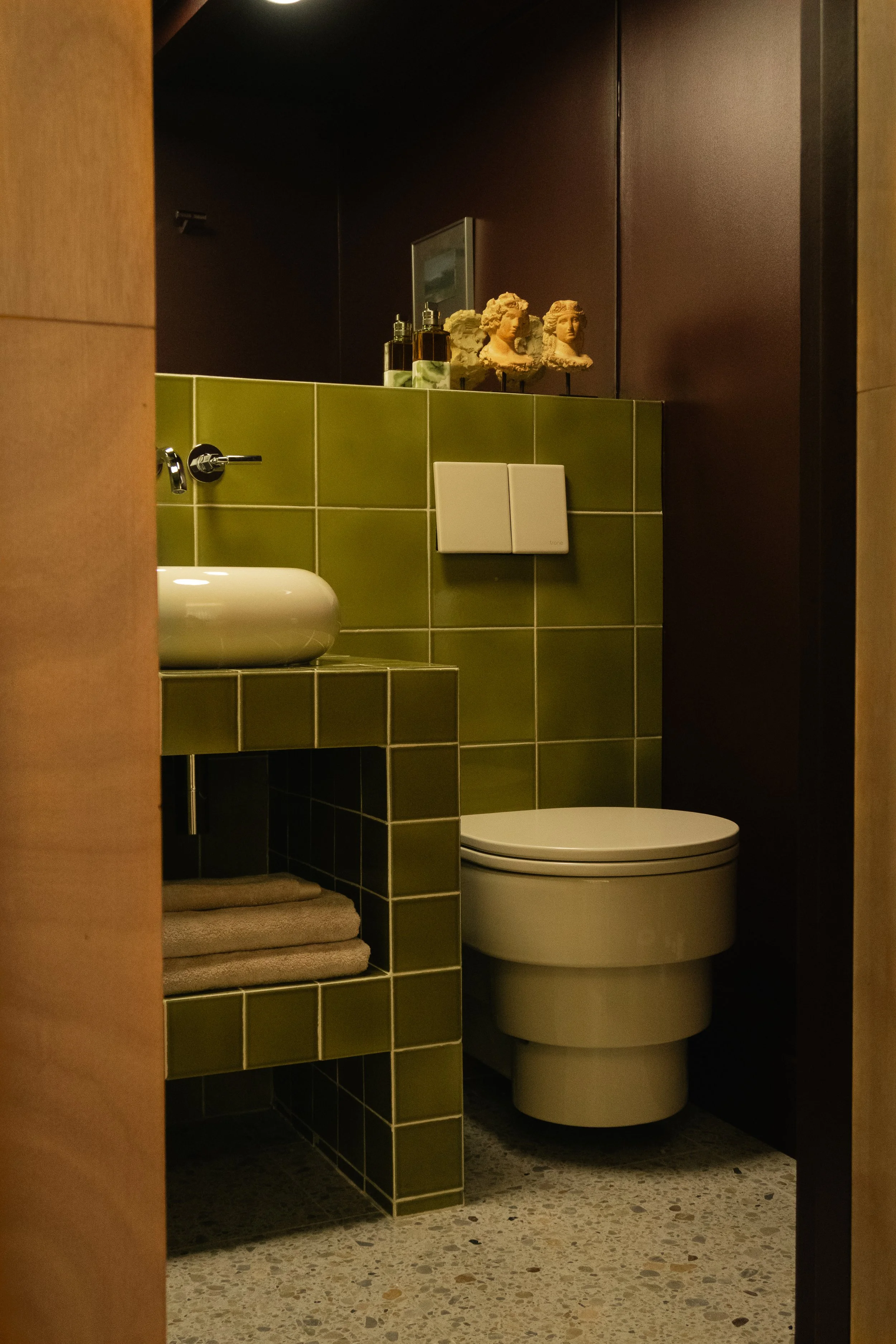 Petite salle de bain avec des carreaux verts, un lavabo blanc, un WC moderne, et des décorations comme des sculptures et des bouteilles sur une étagère. 