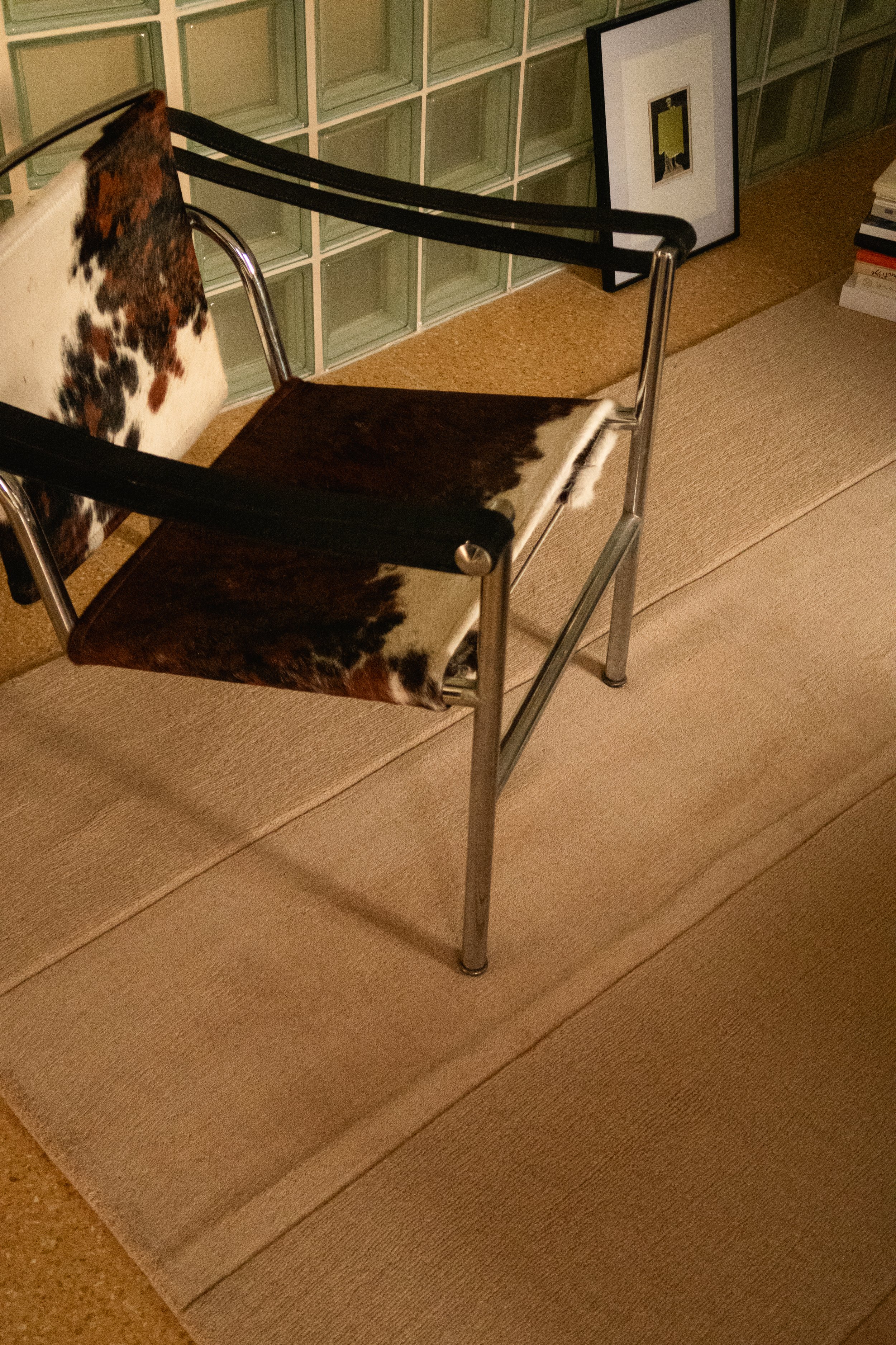 Une chaise avec un siège et un dossier en cuir de vache, en métal chromé, placée sur un tapis beige dans une pièce avec un mur en blocs de verre. Il y a des cadres et des livres à côté.