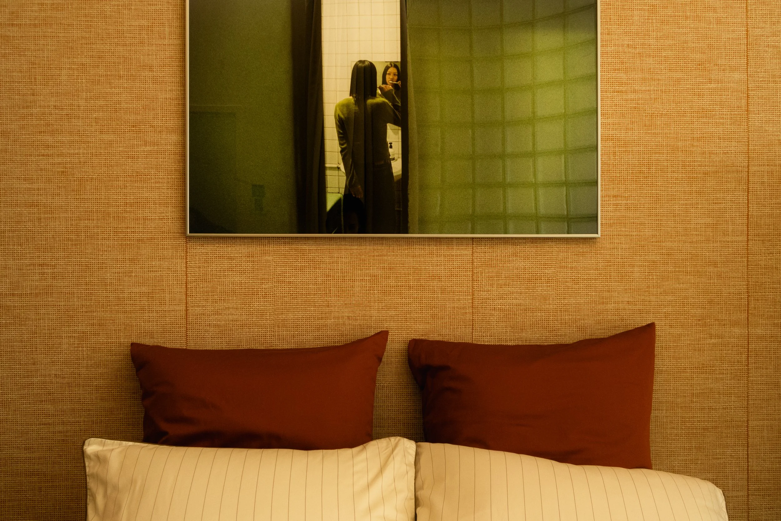 Un miroir au-dessus d'un lit avec deux oreillers marron, dans une chambre avec un mur en revêtement beige et un miroir reflétant une femme prenant une photo.