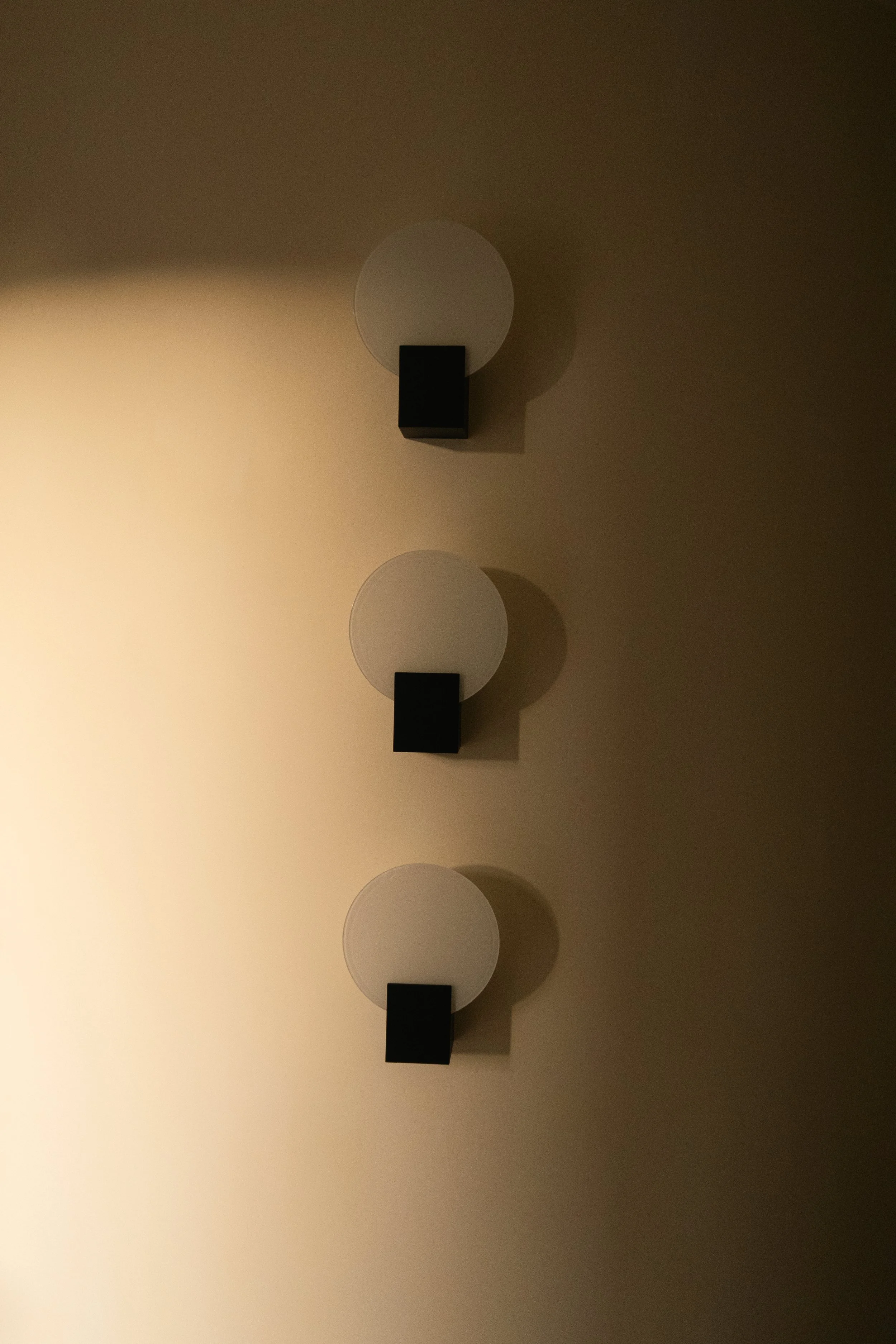 Trois appliques murales avec disque blanc et support noir, alignées verticalement sur un mur beige, avec des ombres portées sur le mur.