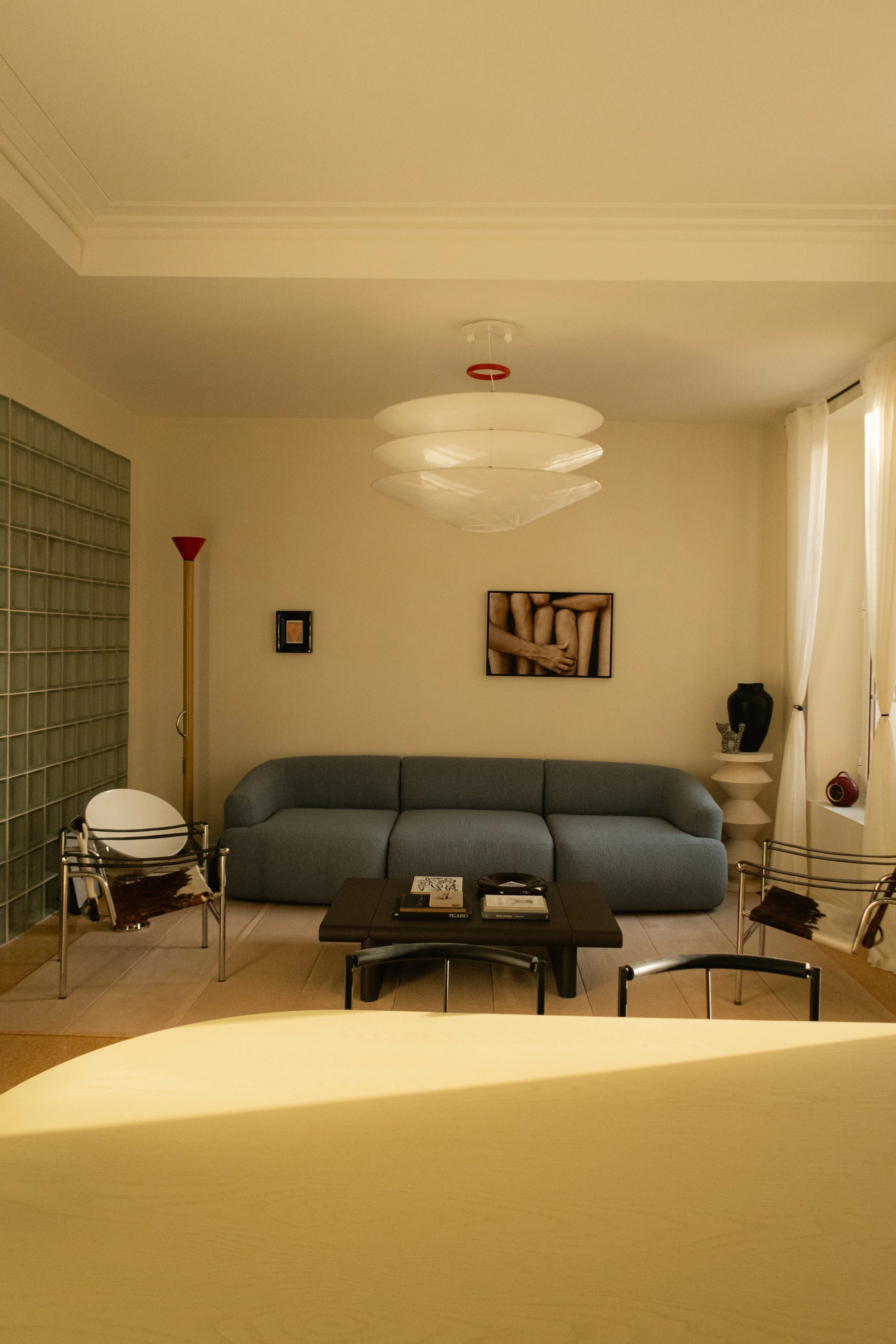 Salon moderne avec canapé gris, deux chaises en métal avec un tapis animal à côté, une table basse en bois, une lampe sur le mur, une peinture, et des rideaux blancs.