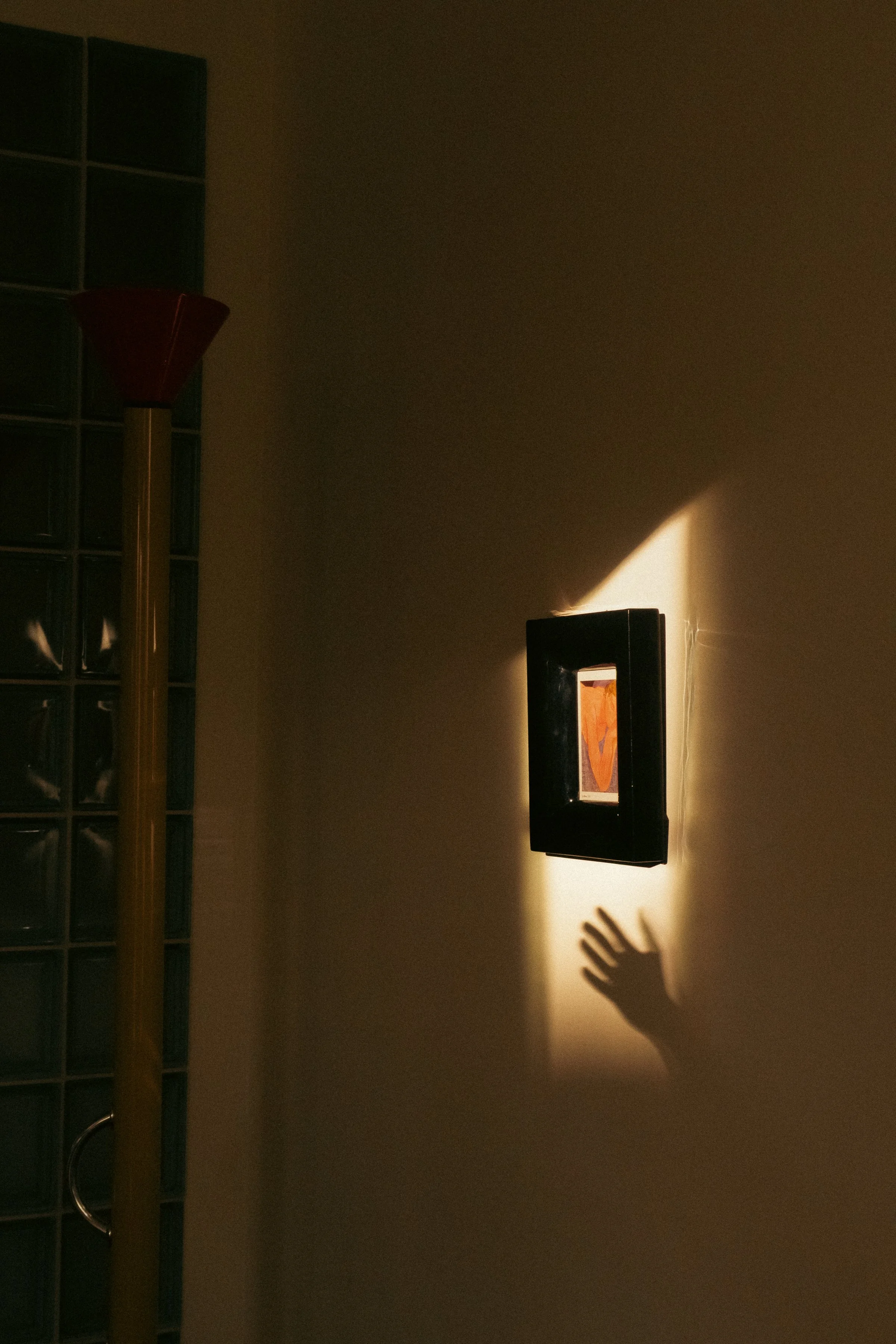 Une pièce sombre avec une lumière qui éclaire partiellement un mur. Sur le mur, il y a un cadre noir avec une image à l'intérieur, et une ombre d'une main est projetée sur le mur, près du cadre.