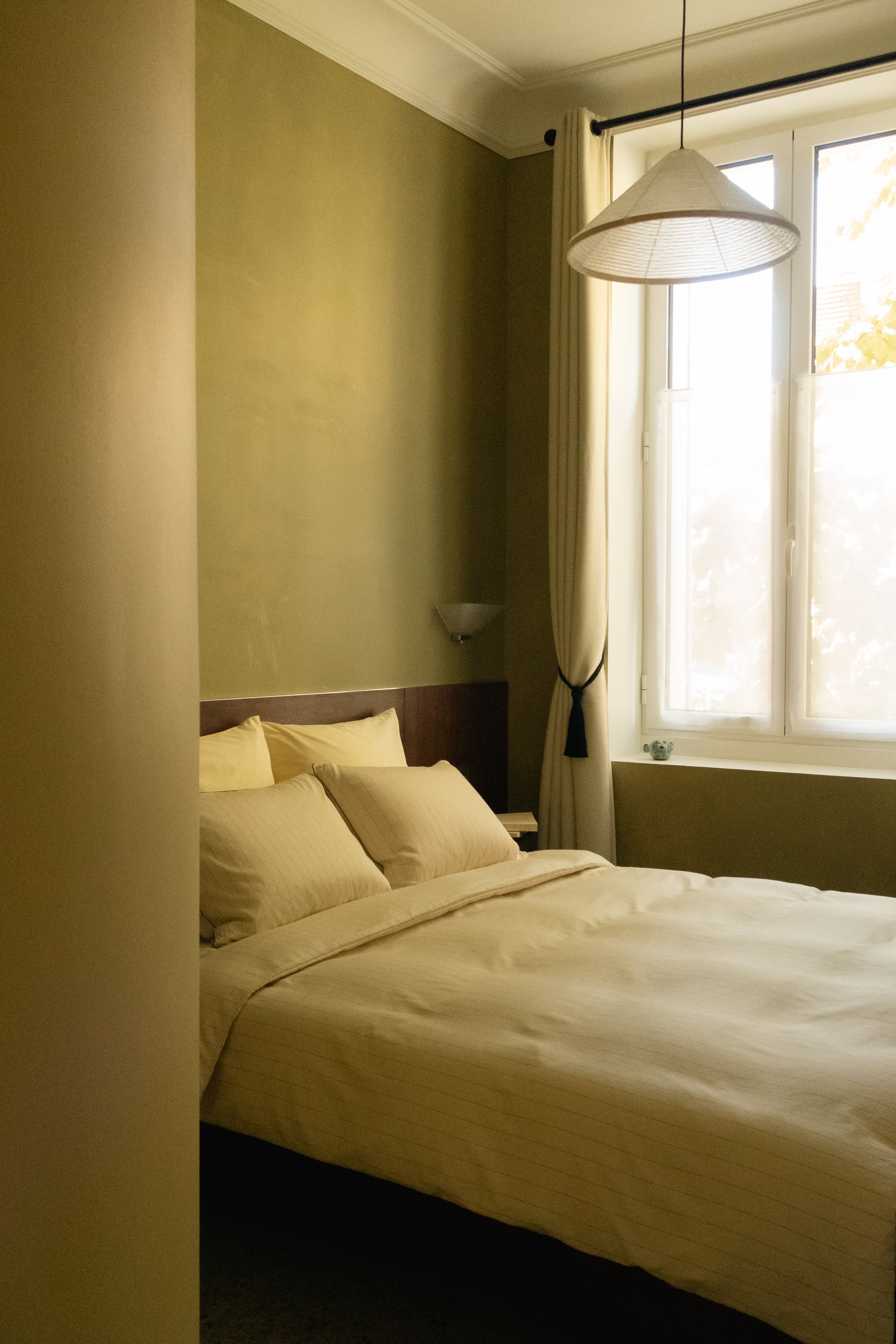 Chambre d'hôtel avec un lit double, des oreillers beiges, une fenêtre avec rideaux, et une lampe suspendue.