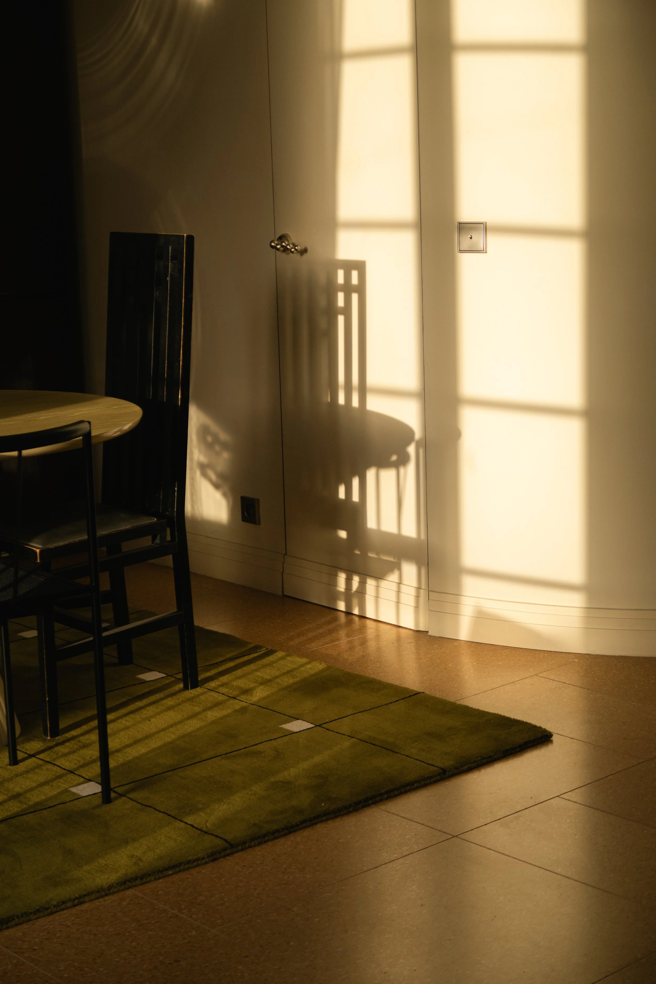 L'ombre d'une chaise sur un mur éclairé par la lumière du soleil à travers une fenêtre. La scène inclut une table et des chaises à côté d'un tapis vert sur un sol en carreaux.