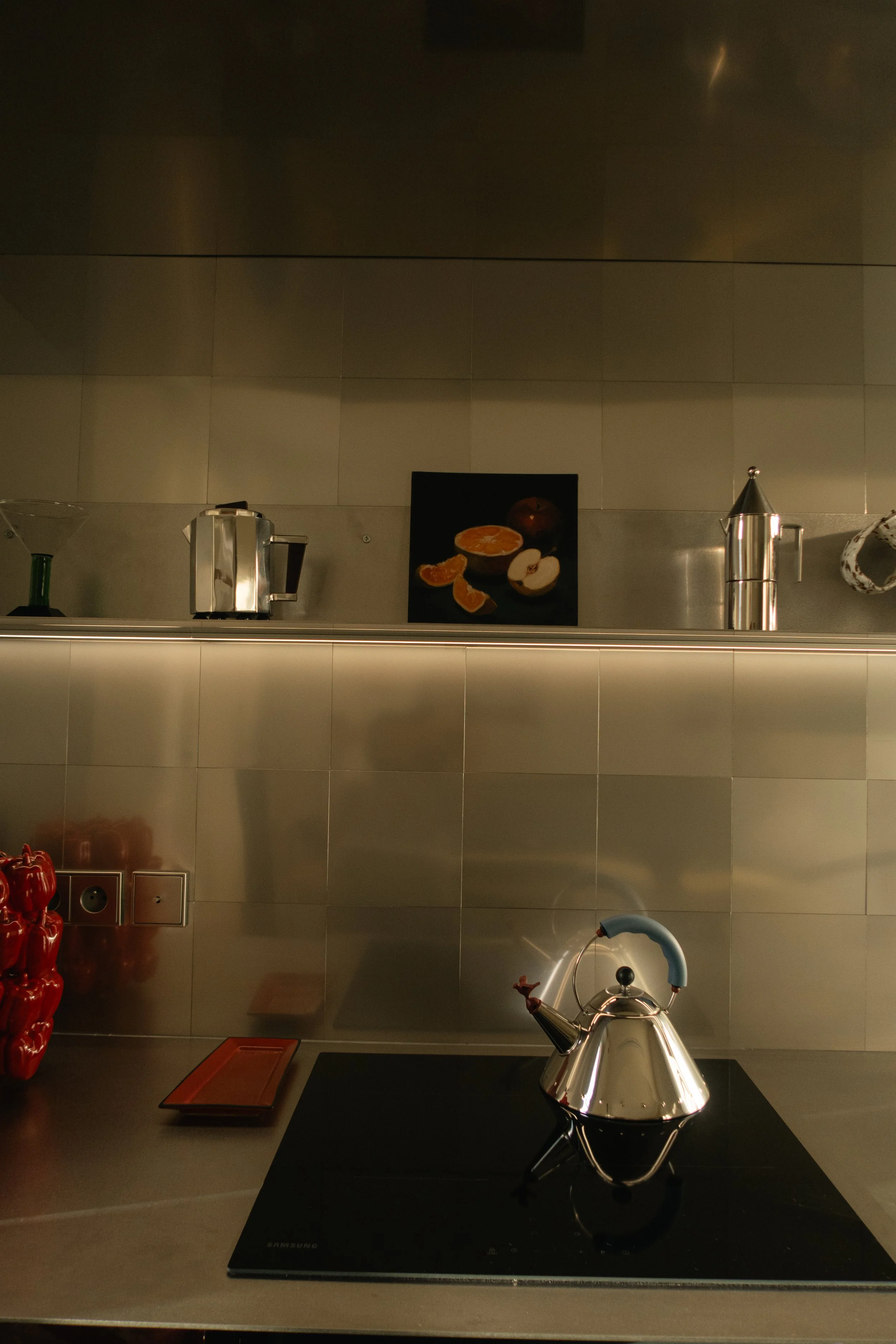 Une cuisine moderne avec une cuisinière à induction, un théière en métal, des appareils électroménagers en acier inoxydable, une peinture représentant des agrumes et des pommes, une pile de poivrons rouges sur le plan de travail.