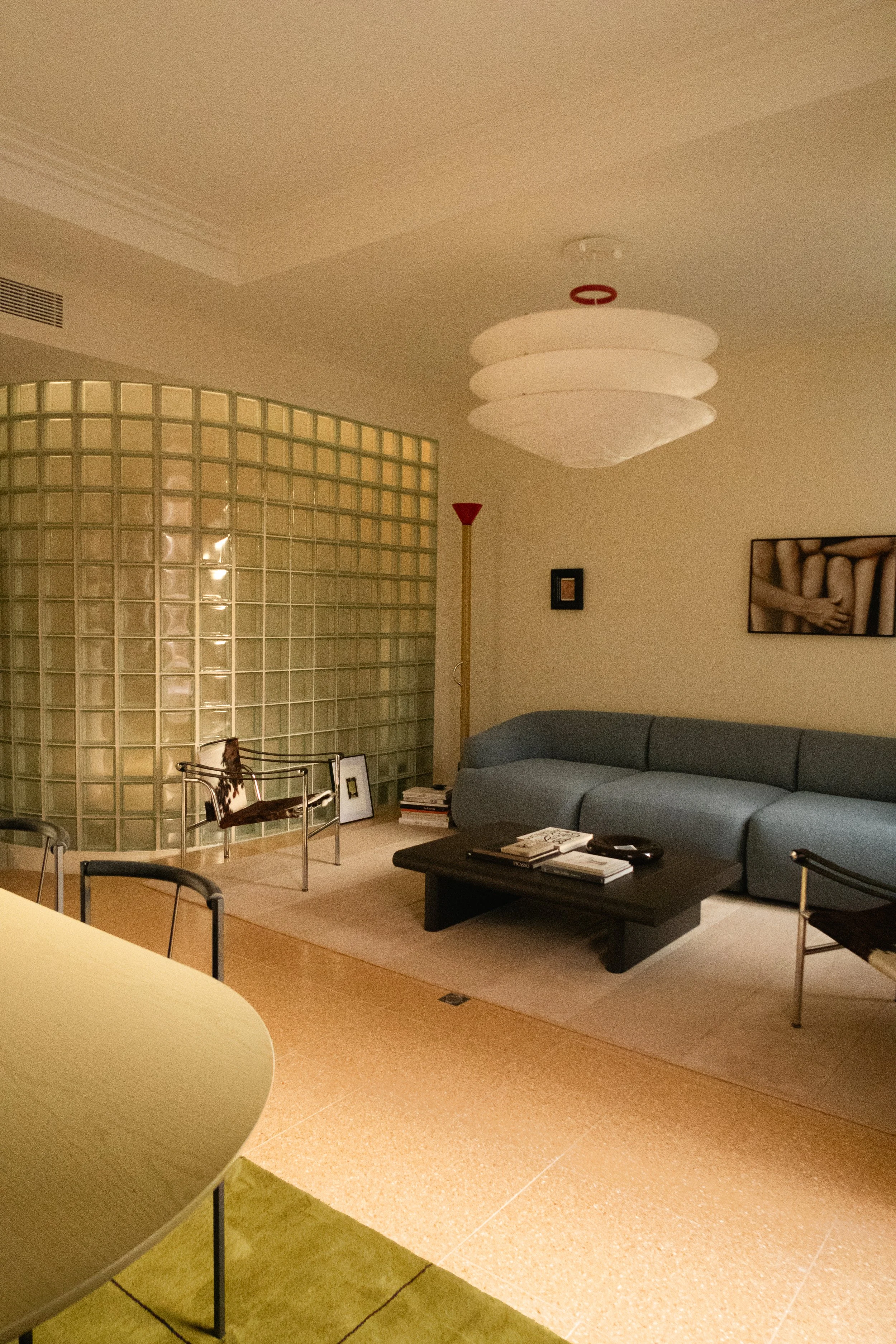 Salon moderne avec canapé gris, table basse, chaise en métal, lampadaire rouge, murs clair, tableau de photos, et paravent en verre.