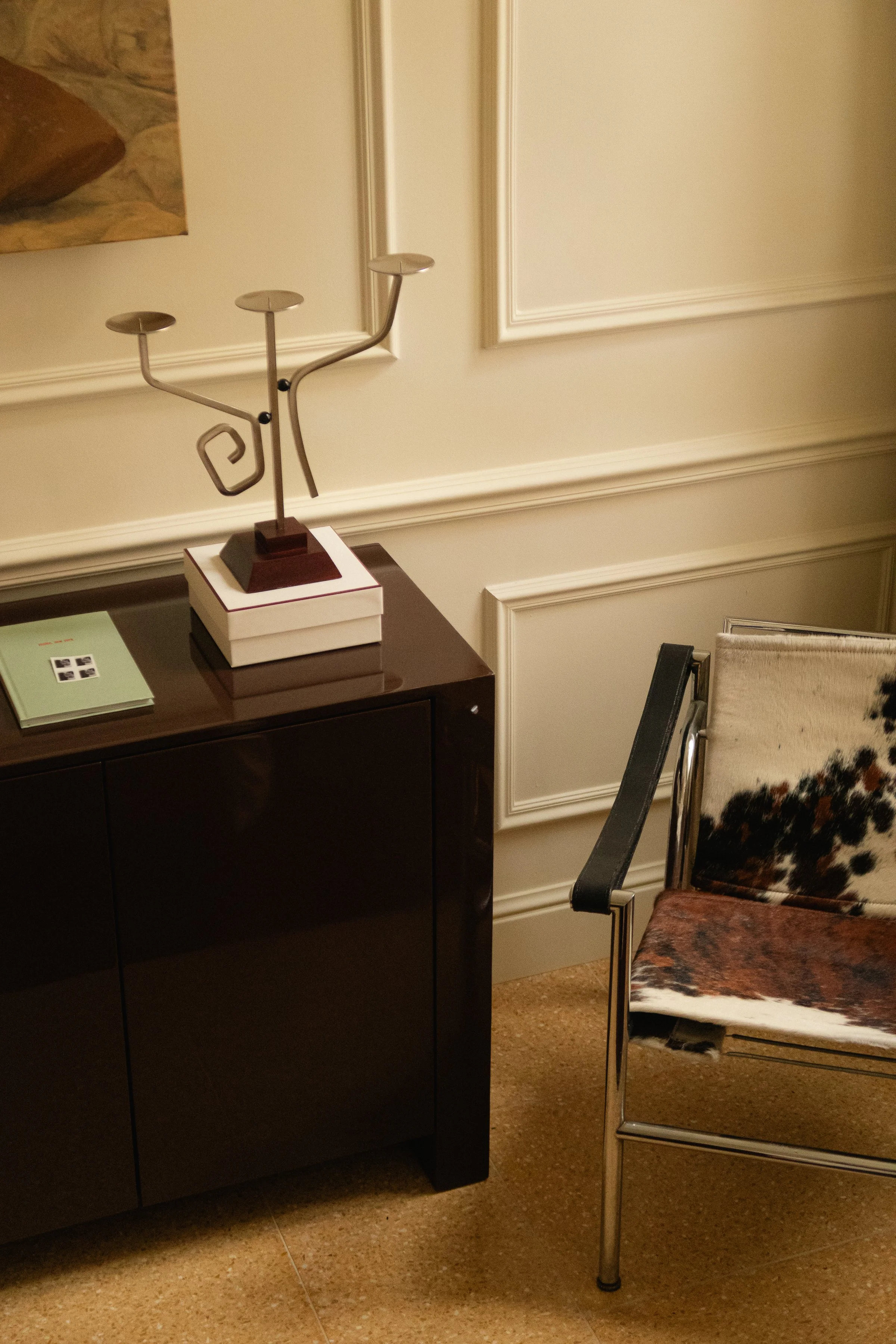 Une console noire avec une lampe en forme de sculpture abstraite dessus, un carnet vert à gauche, et un fauteuil avec un motif animal en cuir, dans une pièce avec des murs crème et des moulures décoratives.