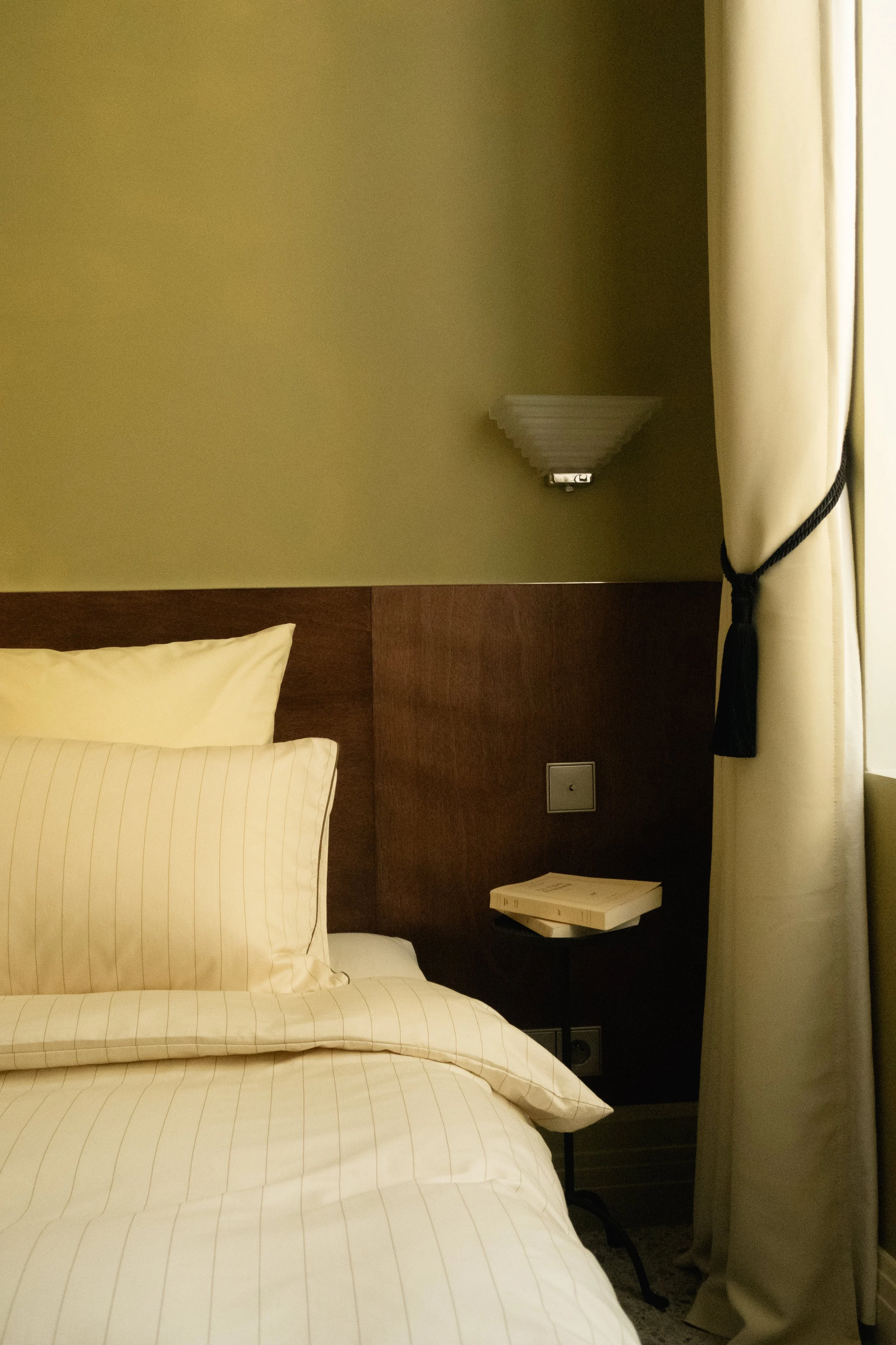 Littérature de lit avec oreillers en drap rayé crème et beige, lampe murale, table de nuit avec livres, et rideau beige à nœud noir dans une chambre d'hôtel.