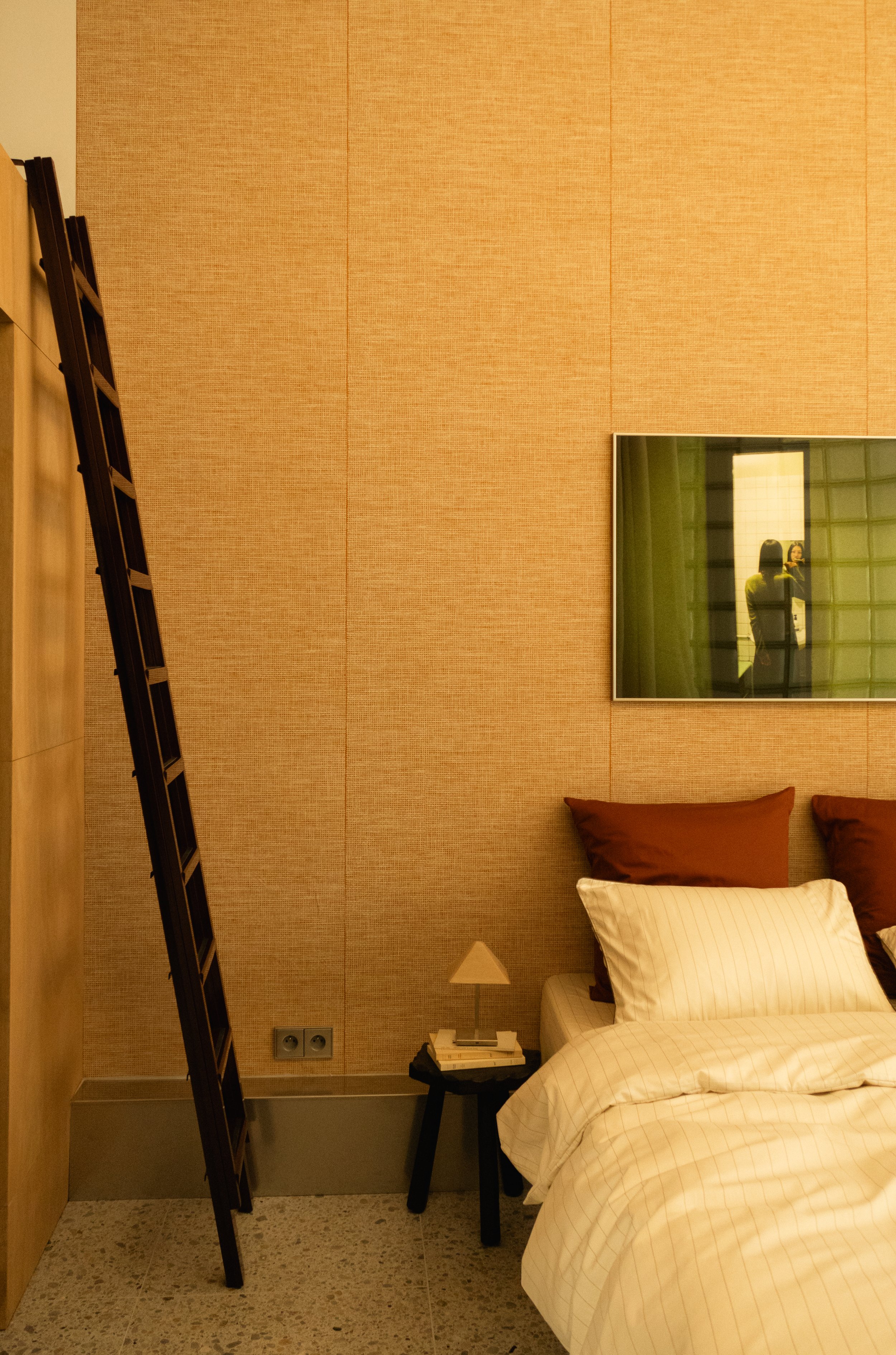 Intérieur d'une chambre avec un lit, deux oreillers marron, un oreiller blanc rayé, une table de chevet avec une lampe et un téléphone, un miroir et un tableau sur le mur, et une échelle noire en bois