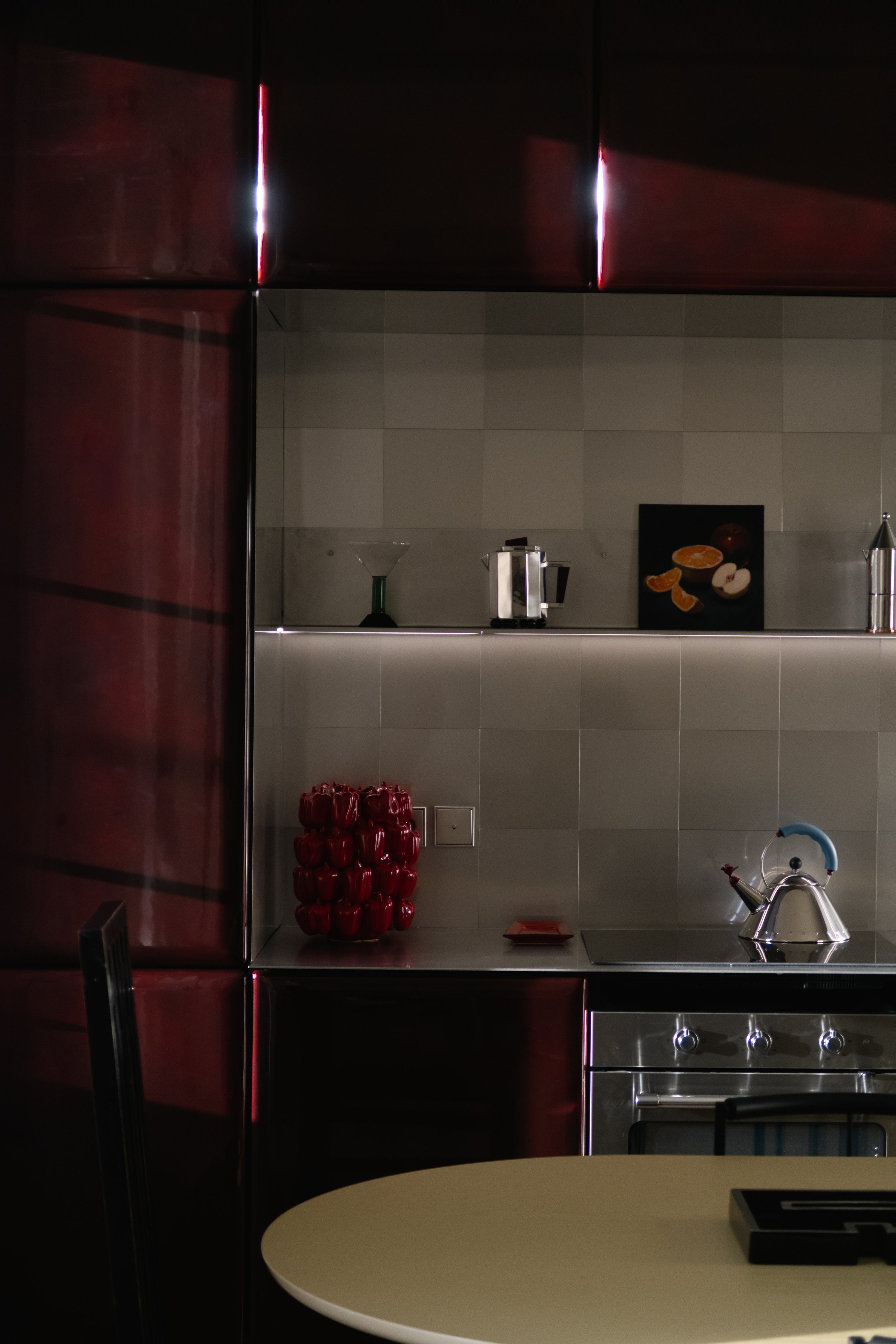 Une cuisine moderne avec des surfaces en métal, un four, une bouilloire, un tableau avec des fruits et des appareils de cuisine sur une étagère, et une table ronde beige partiellement visible au premier plan.