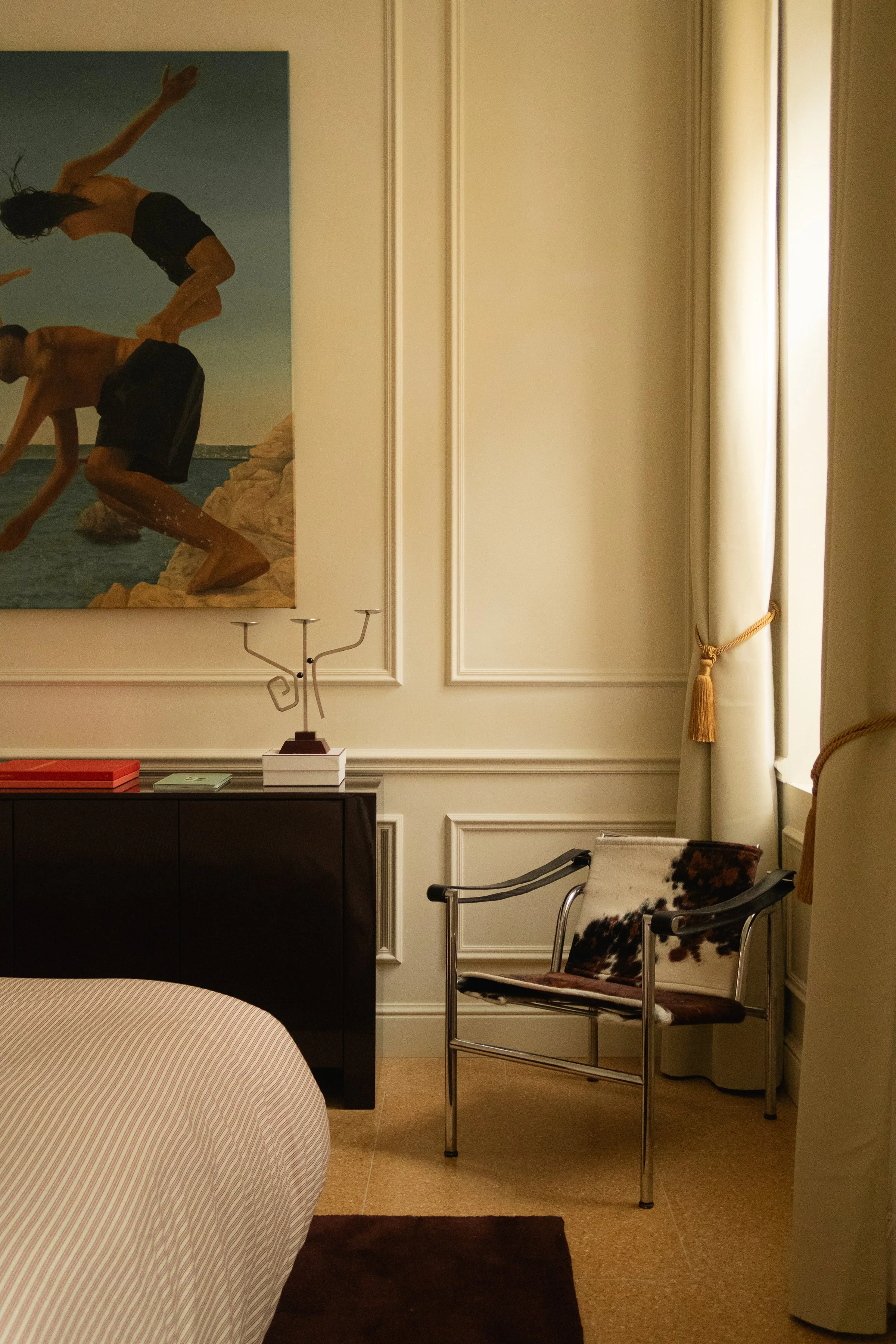 Intérieur d'une chambre avec un fauteuil en cuir et une porte de fenêtre avec rideau beige, mur blanc avec moulures, et un tableau représentant des personnes pliant sur la plage.