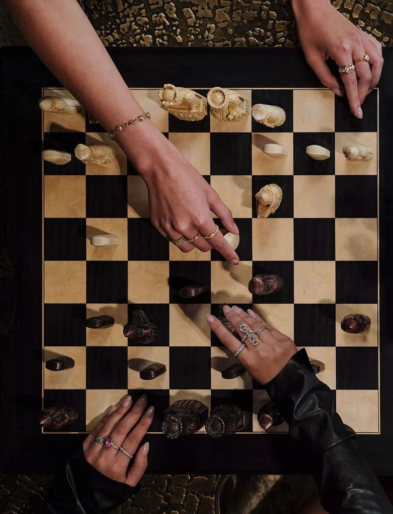Deux personnes jouent aux échecs, une personne avec des ongles longs noirs et des bijoux dorés, l'autre avec des bijoux argentés et des vêtements en cuir noir, en train de déplacer des pièces en bois sur un échiquier en bois clair et foncé.
