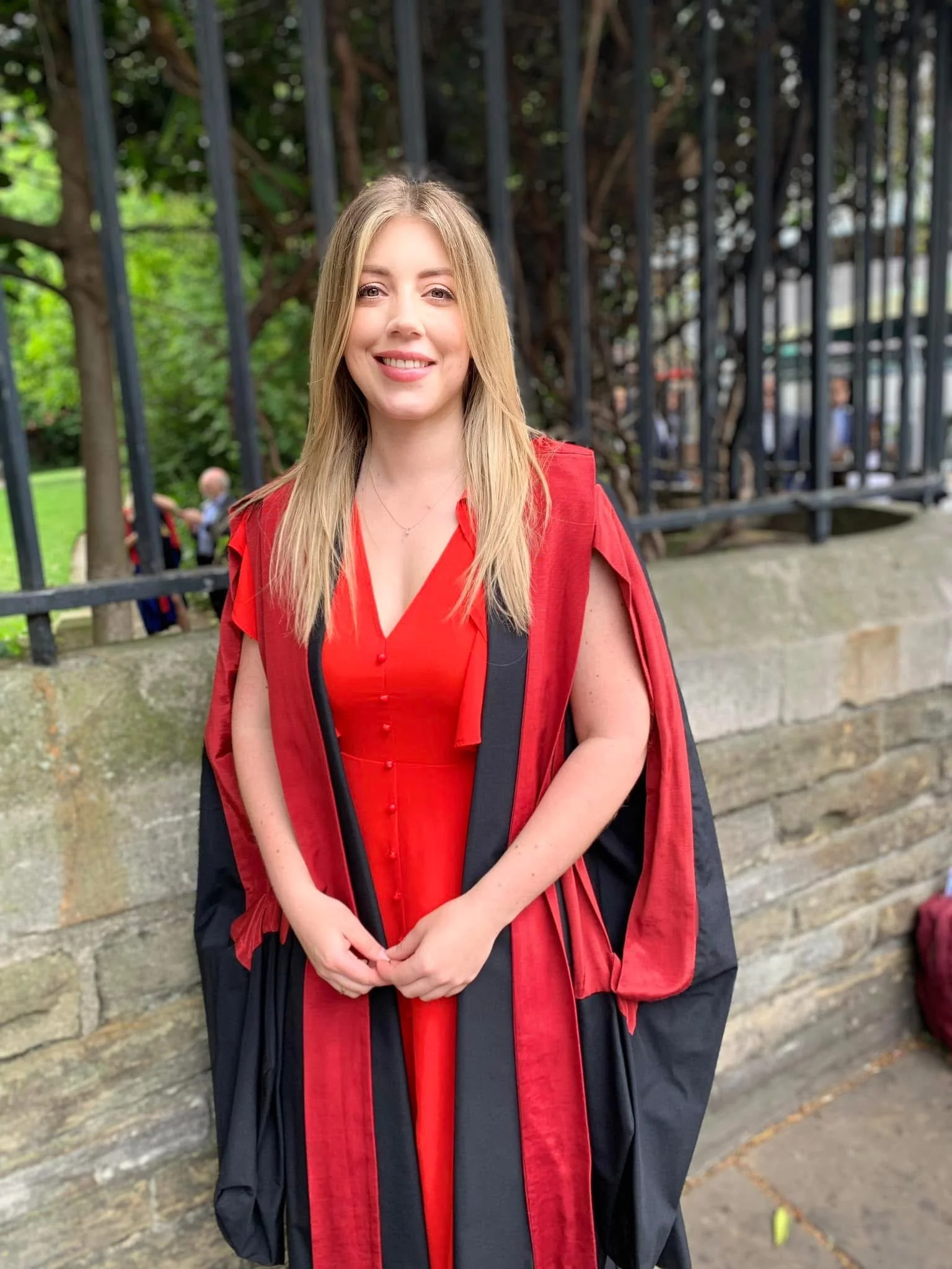 A woman with blonde hair in a red dress and black academic gown and mortarboard cap, smiling, standing outdoors with trees and a brick wall in the background.