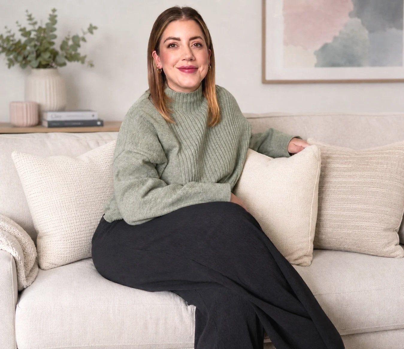 A woman with light brown hair sitting on a light-colored couch with beige cushions, wearing a green turtleneck sweater and black pants, smiling and looking at the camera in a cozy living room.
