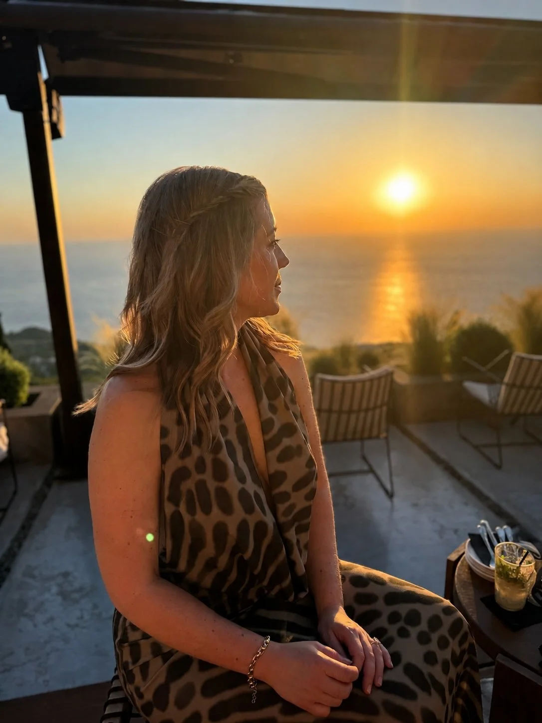 A woman sitting on a patio during sunset with the ocean in the background, wearing a leopard print dress, with shoulder-length wavy hair, and a bracelet on her wrist.