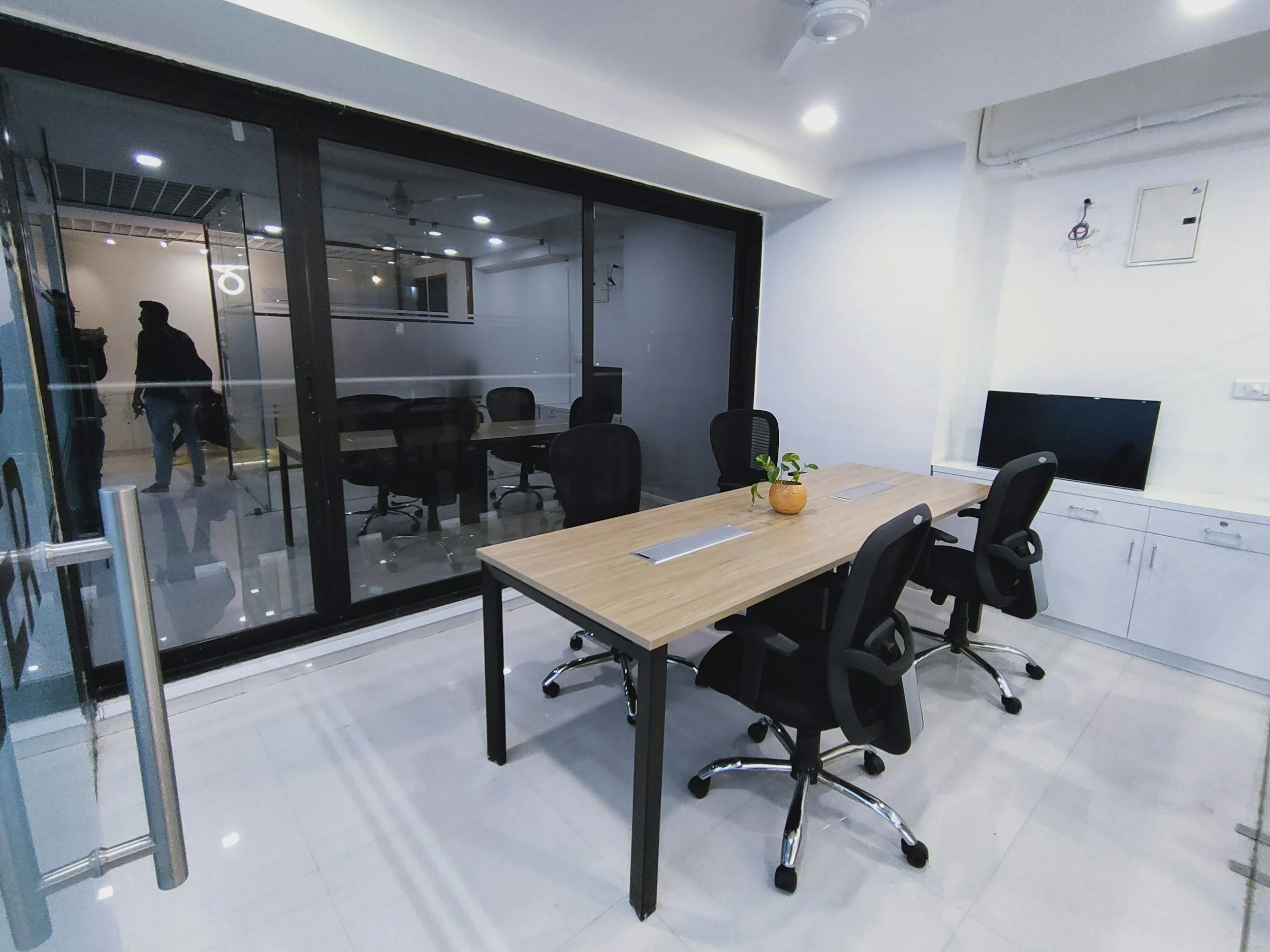 Modern office meeting room with a wooden table, four black office chairs, a small potted plant, and a television on white cabinet, separated from another meeting room by a glass partition.