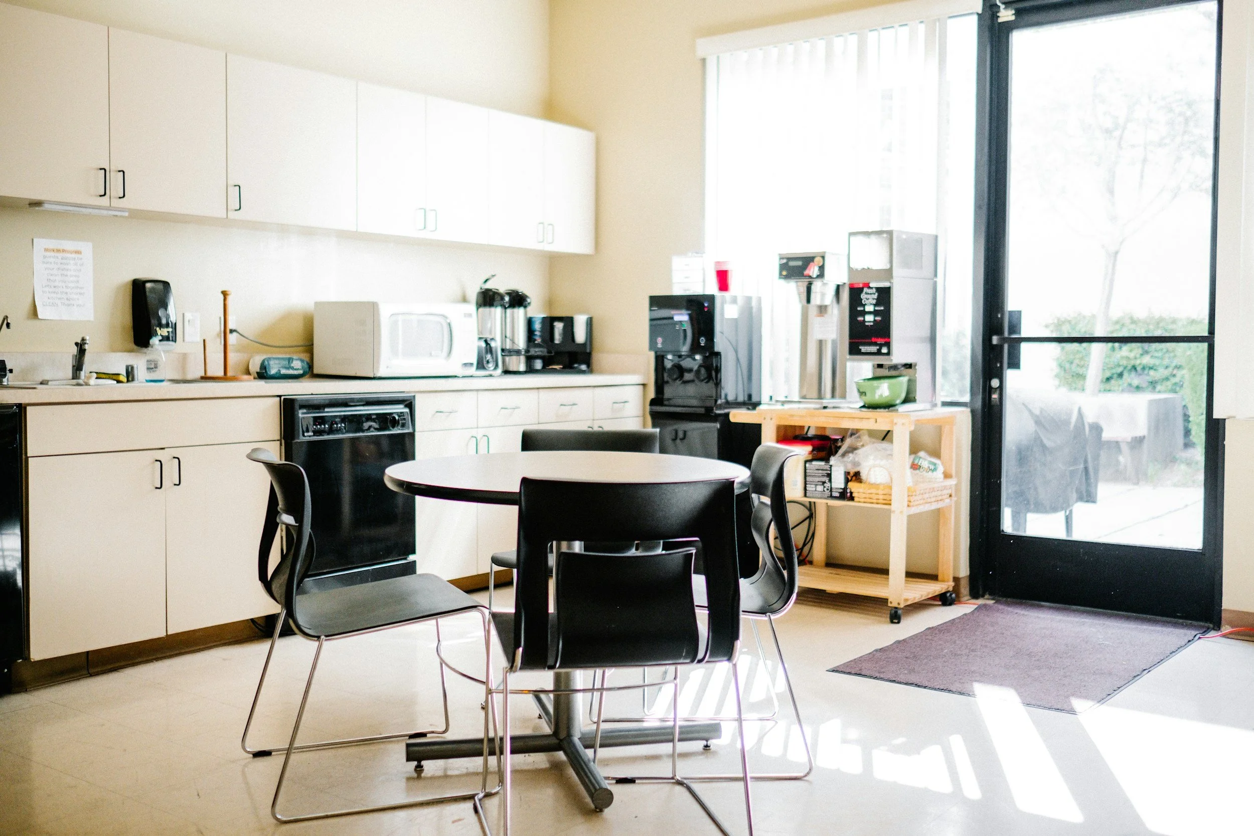Kitchen area with black table and chairs, microwave, coffee machine, and water dispenser near a glass door.